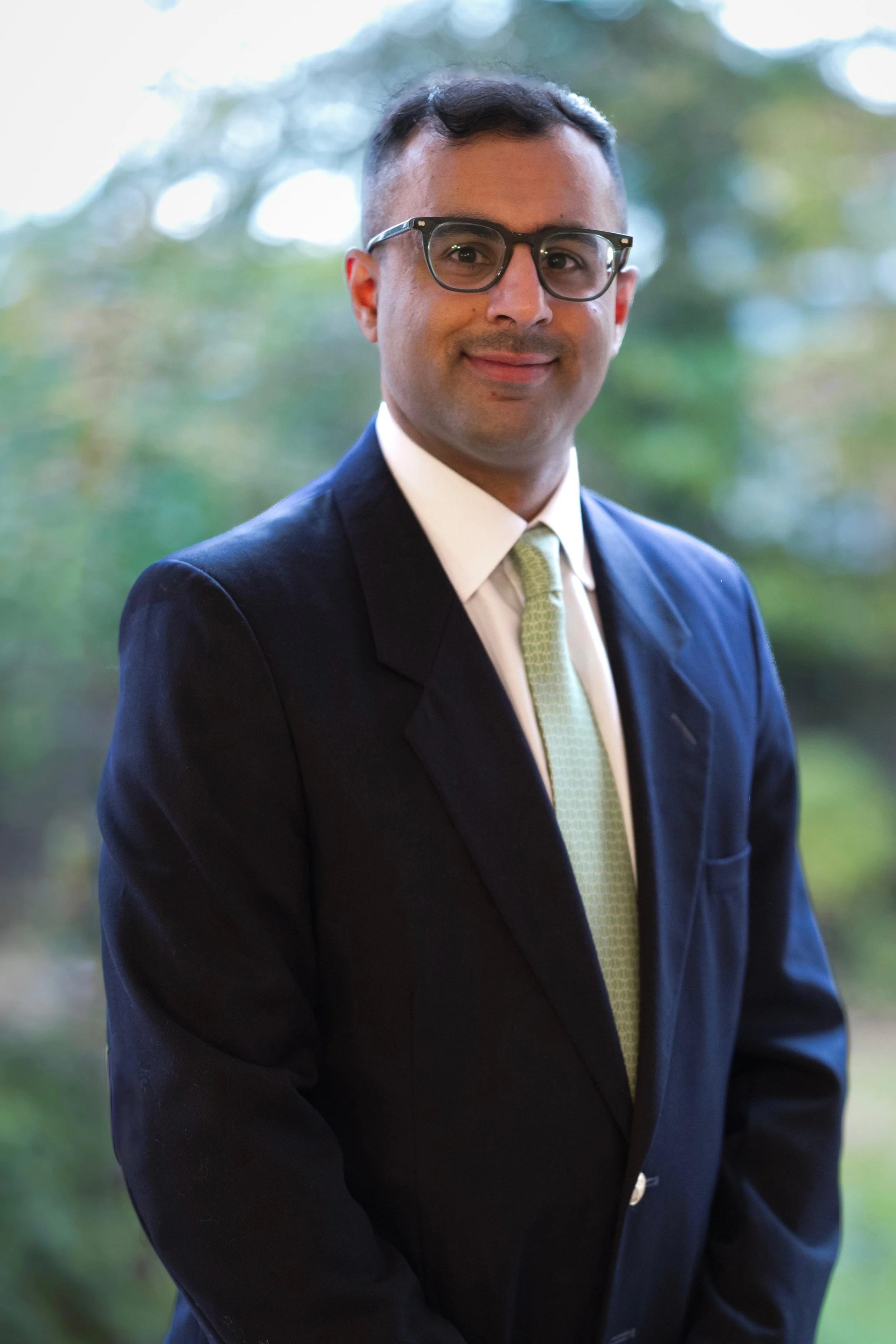A male doctor with glasses, dark hair and a beard, wearing a white coat and blue shirt, standing with arms crossed against a plain background, with a stethoscope around his neck.