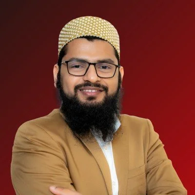 A man wearing a beige traditional hat, glasses, and a tan blazer smiling against a red background.