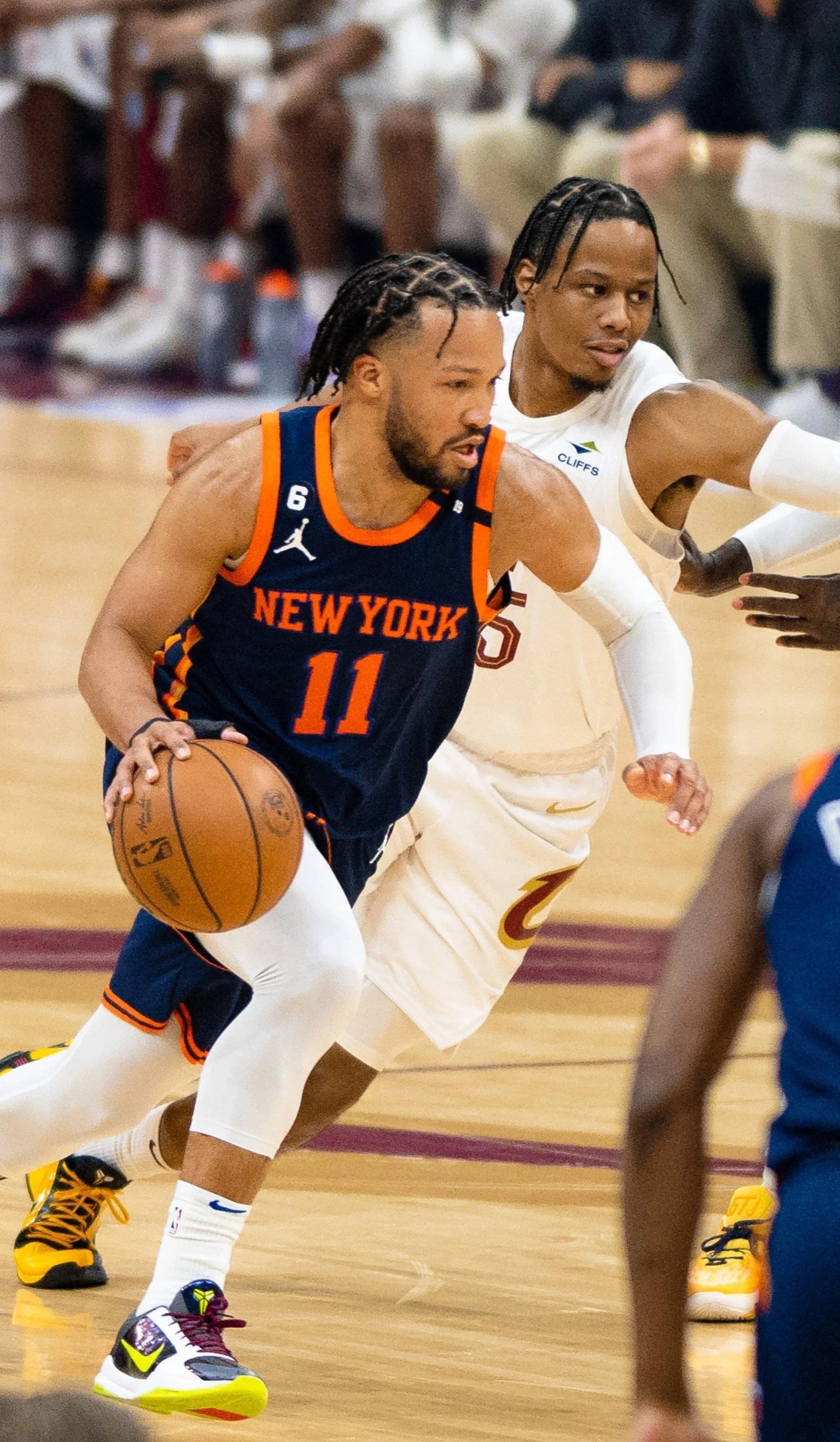 A basketball game with players in motion, one player from New York in a navy jersey with orange accents dribbling the ball, and another player from Cleveland Cavaliers defending.
