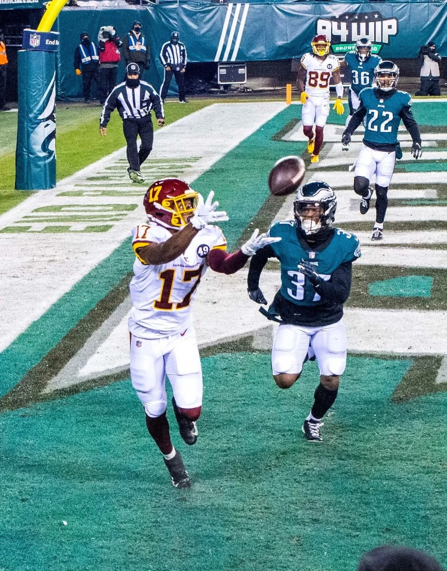 Two football players, one in a white jersey number 17 and another in a darker jersey number 3, running toward the end zone where a football is about to cross the goal line. A referee is in the background, along with other players and officials on the field.