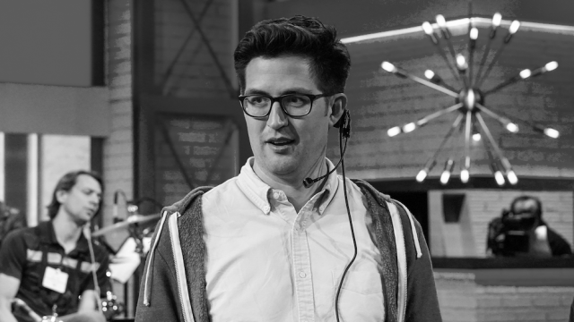 Man wearing glasses and a headset on a film or TV set with crew members and colorful lighting in the background.