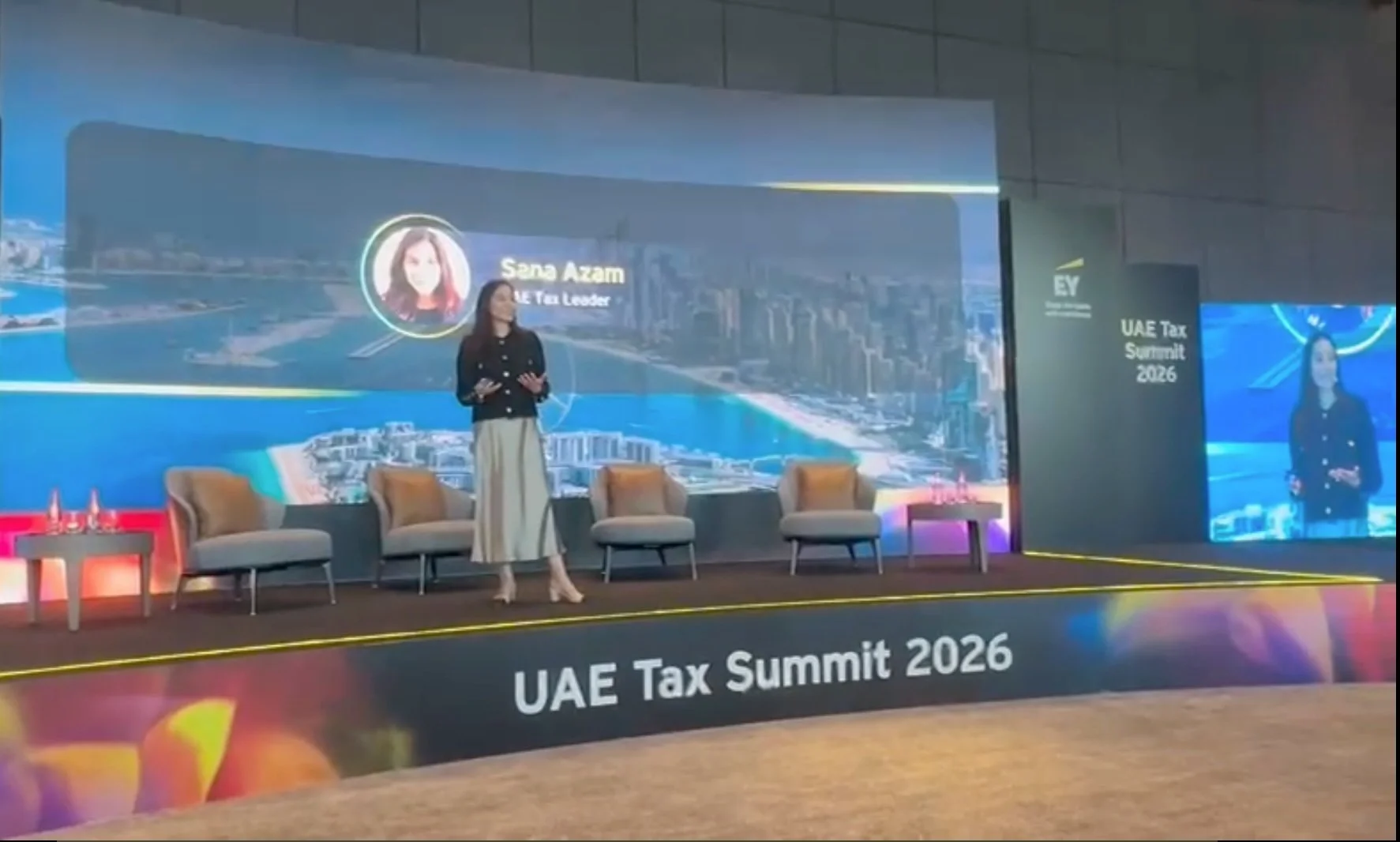 A woman speaker standing on a stage at the UAE Tax Summit 2026, with a large screen behind her displaying her profile picture, name 'Sana Azam', and the event title 'UAE Tax Summit 2026'. There are three beige chairs on stage and a small side table with water bottles and glasses.