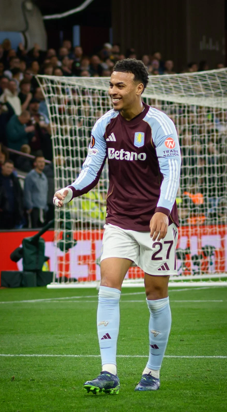 A soccer player in a maroon and light blue jersey with the number 27 on his shorts, smiling and celebrating on the field during a match.