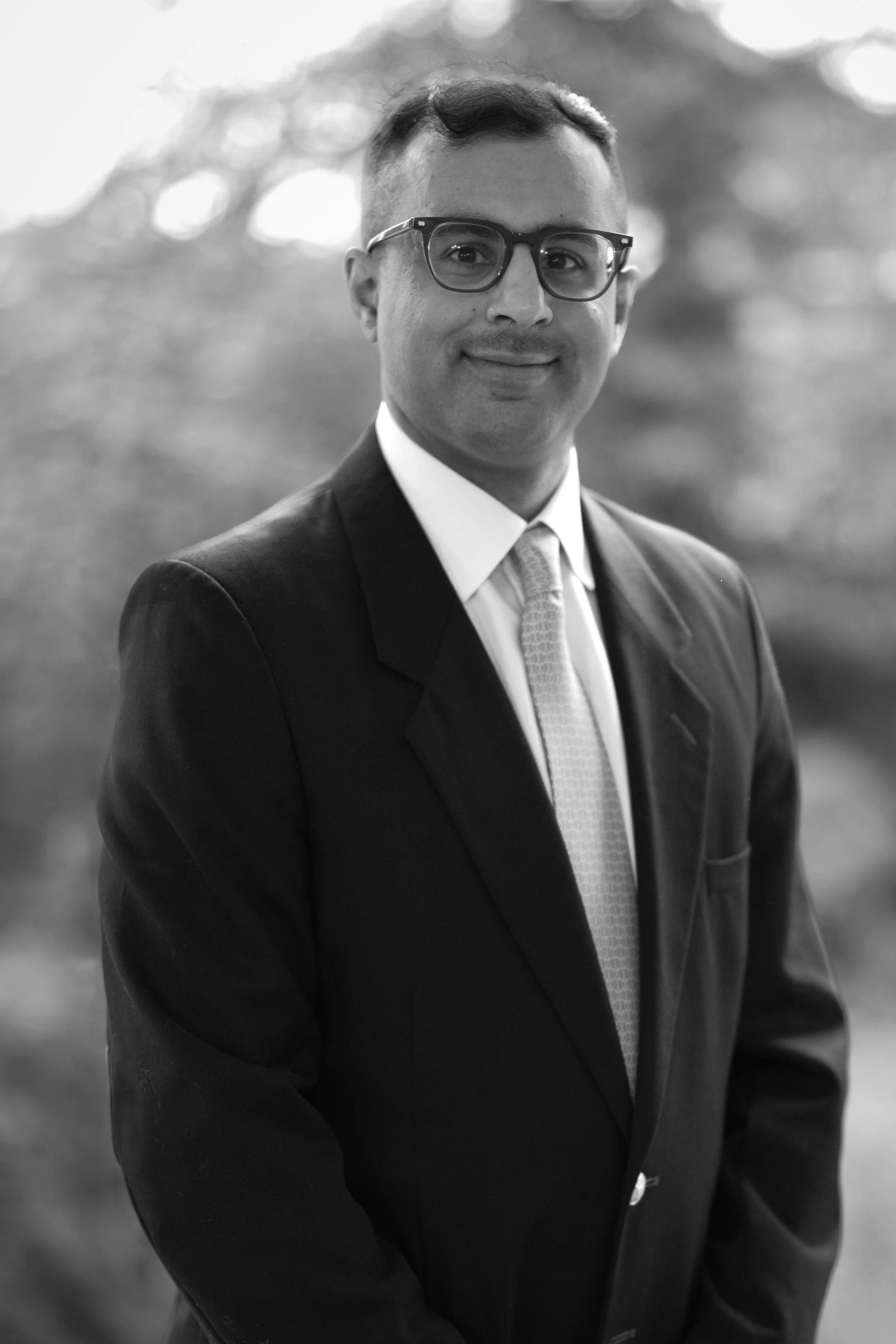 A male doctor with glasses, dark hair and a beard, wearing a white coat and blue shirt, standing with arms crossed against a plain background, with a stethoscope around his neck.
