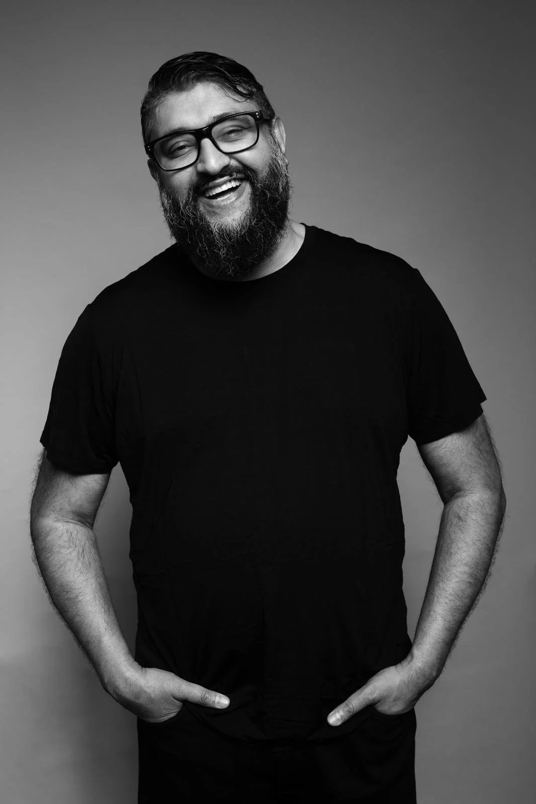 A smiling man with a beard, wearing glasses and a black t-shirt, standing against a plain gray background.