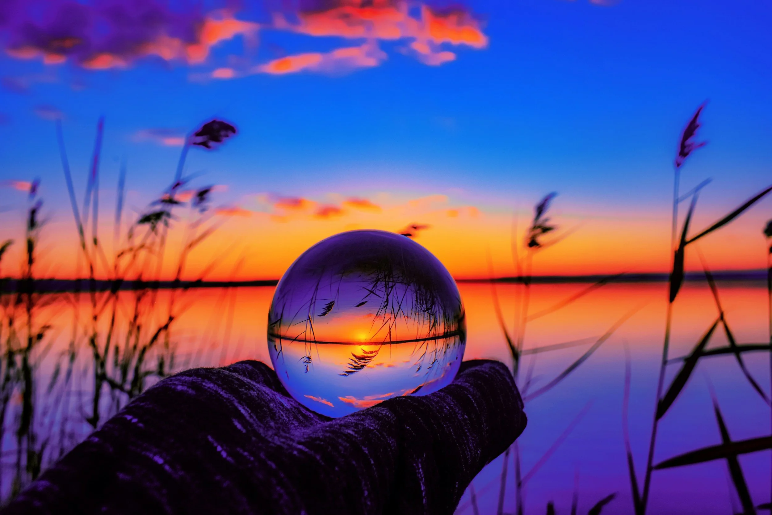 A glass sphere held in a person's hand reflecting a sunset over a body of water with grass in the foreground.