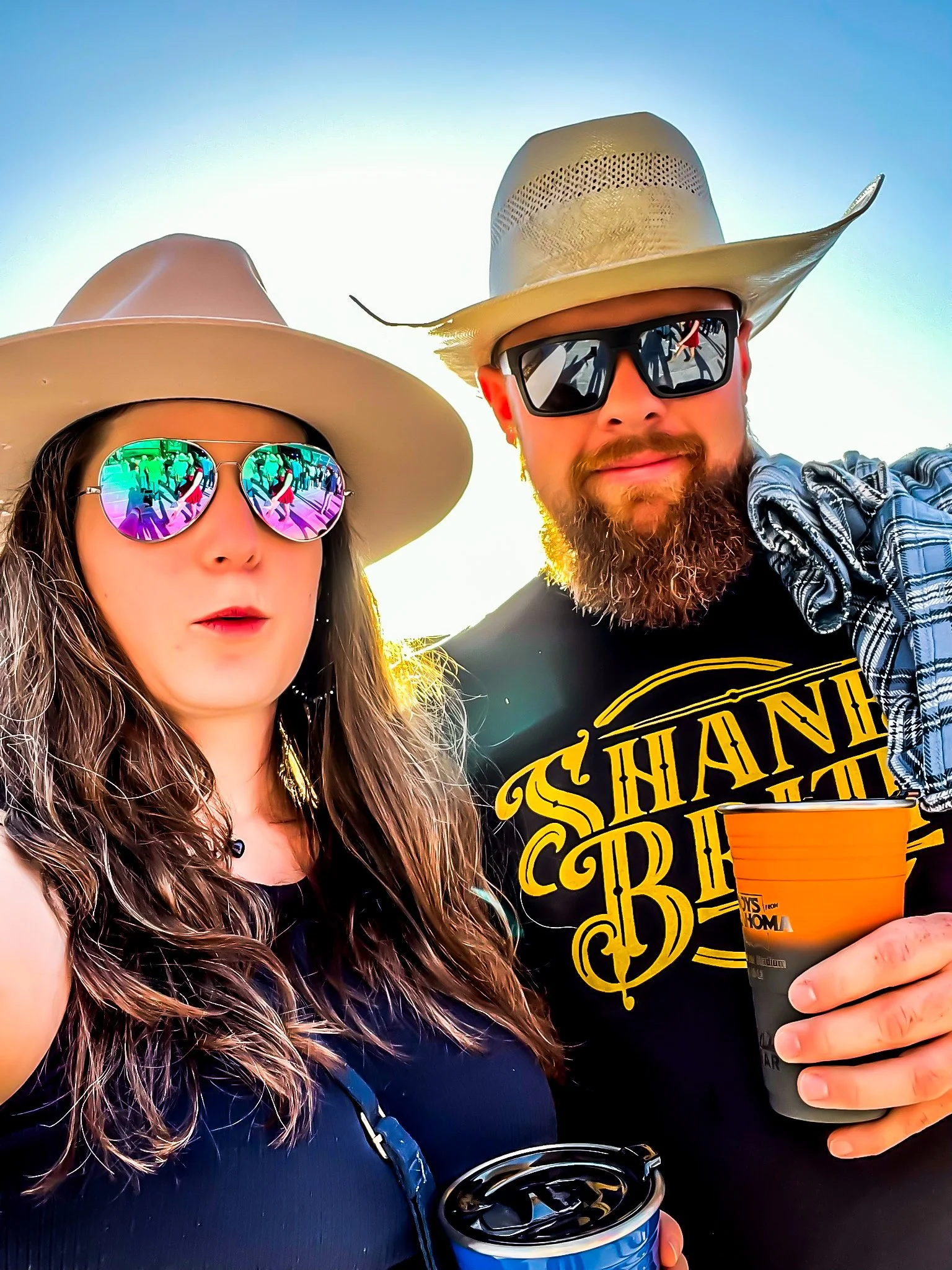 Close-up of a woman with long wavy hair wearing a large beige hat and reflective sunglasses, standing next to a man with a beard, wearing a beige cowboy hat and black sunglasses, holding a black cup, outdoors on a sunny day.