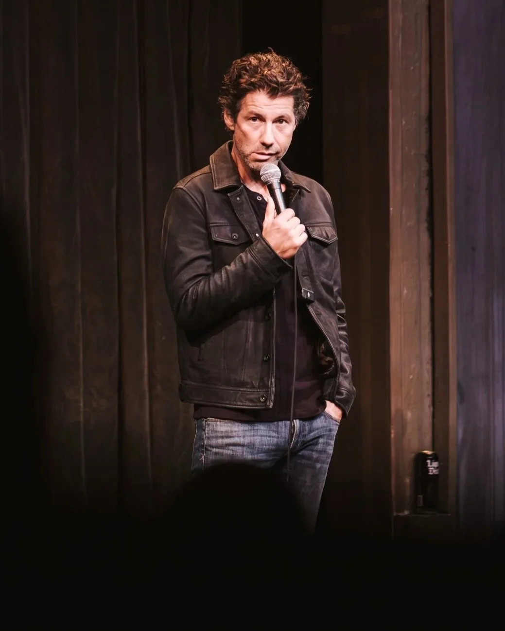 A man with curly hair wearing a black leather jacket and jeans, holding a microphone, performing on stage with dark curtains in the background.
