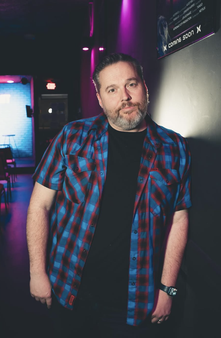 A man with a beard and mustache leaning against a wall in a dimly lit venue with purple and blue lighting, wearing a red and blue plaid shirt and a black t-shirt, with a watch on his left wrist.