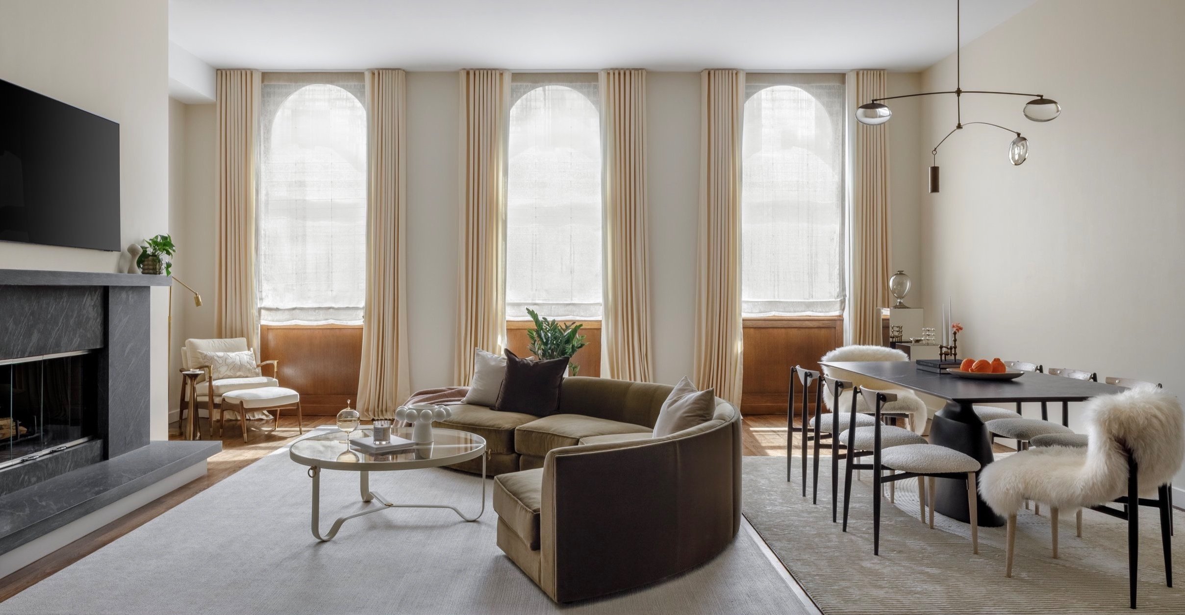 Modern Soho loft living and dining area with curved velvet sofa, marble fireplace, arched wood-framed windows, and minimalist black dining table. Interior design by Studio Kravt, NYC.