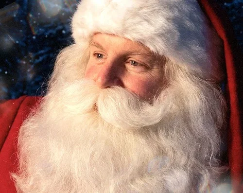 Santa Claus with a white beard and mustache, wearing a red coat and a furry white hat, looking off to the side against a dark, snowy background.