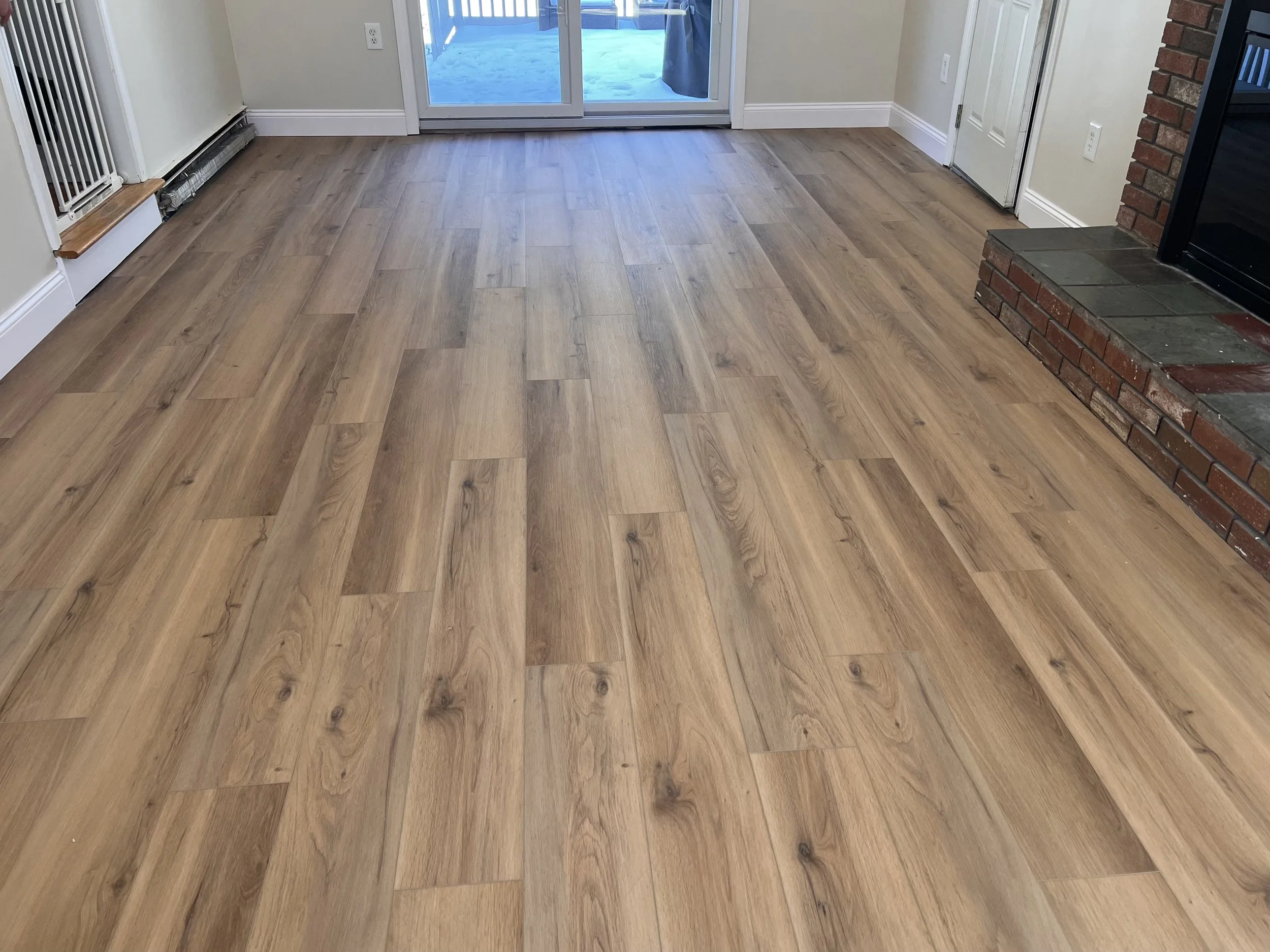 Open-concept living room and kitchen with wooden floors, white walls, and a stone fireplace. The kitchen has white cabinets, a blue island, and clutter on the countertop. Living room has a beige sofa with pillows, a small wooden side table, and large windows.