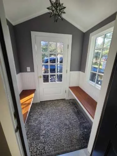 Small entryway with gray walls, two wooden benches on either side, a door with glass panes, and a window on the right, with a patterned rug on the floor.