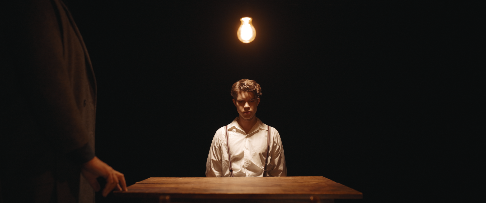 Young man with rolled-up sleeves and suspenders sitting at a wooden table in a dark room illuminated by a single hanging light bulb.