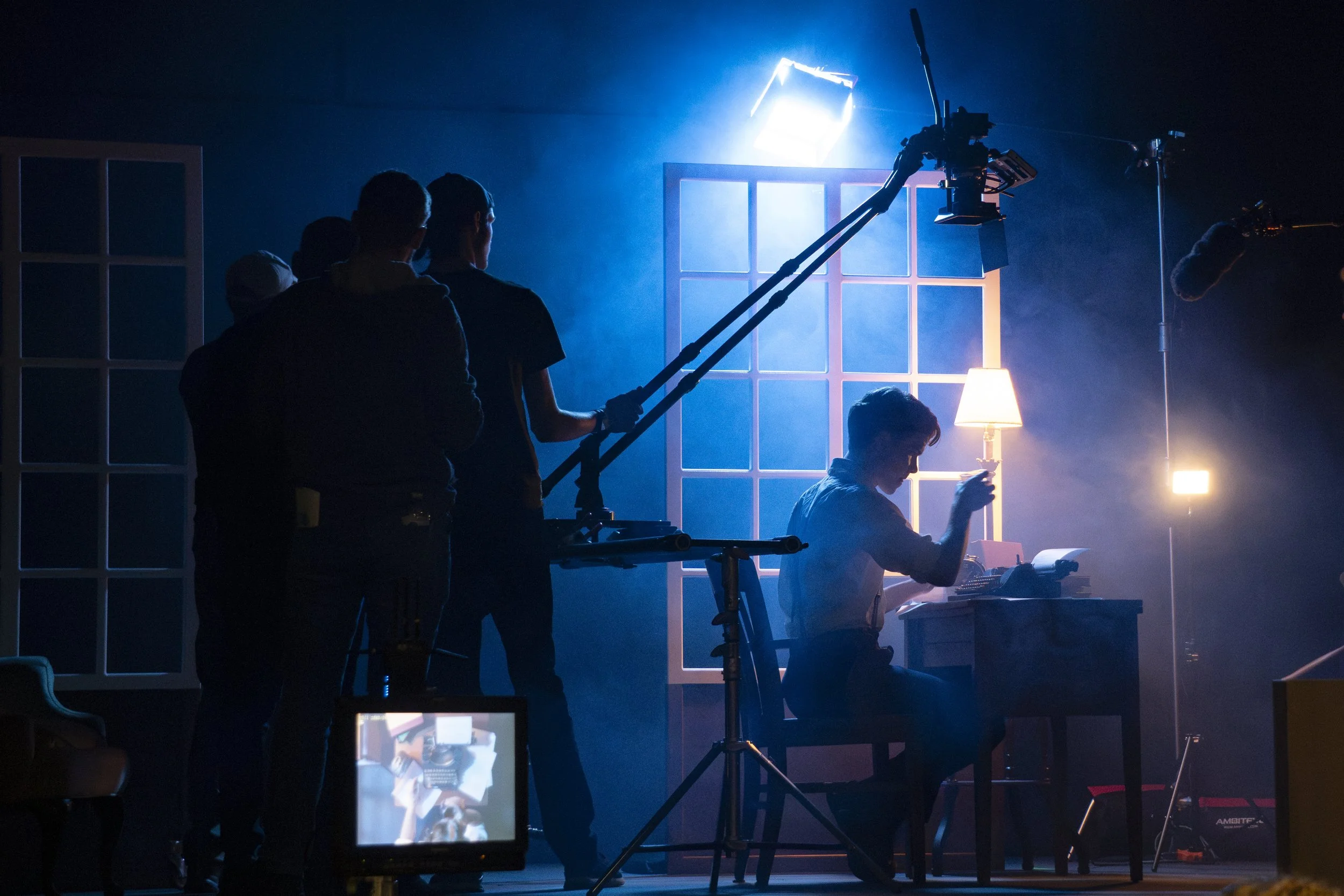 Film crew filming a scene in a dimly lit indoor setting with vintage decor, blue lighting, and a person sitting at a desk using a typewriter.