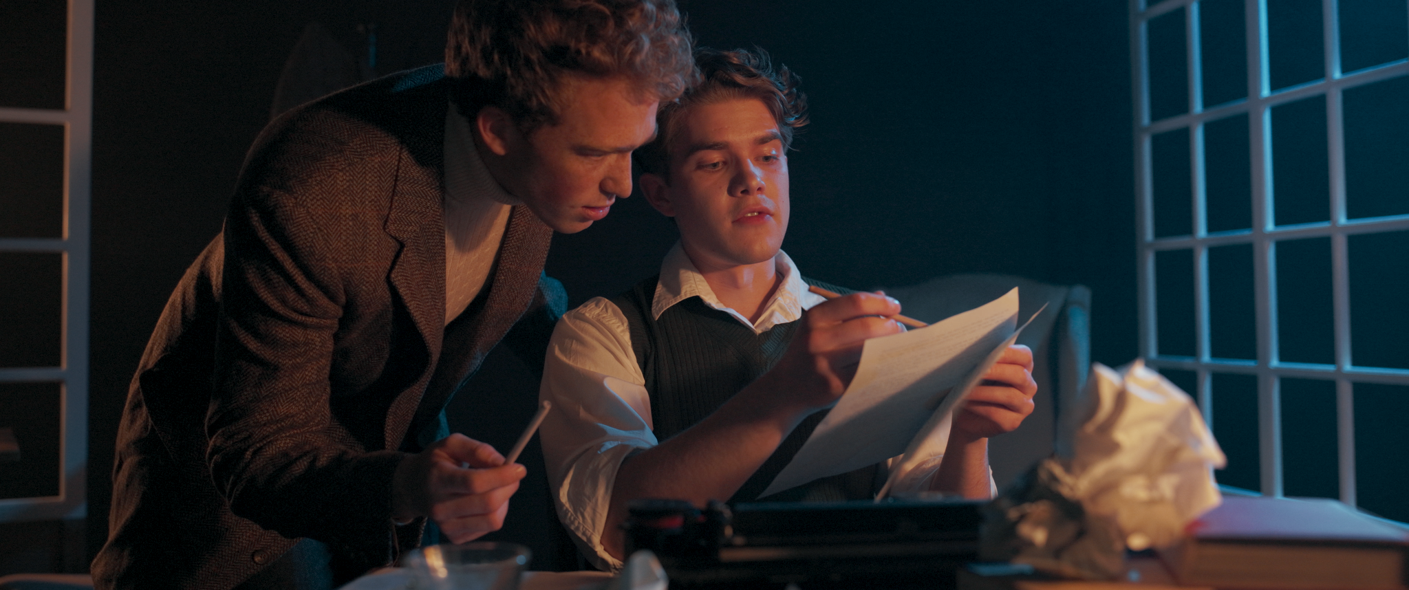 Two young men are inside a dimly lit room with a grid-patterned window. One is sitting on a couch, holding a paper and a pencil, while the other is leaning over him, looking at the paper.