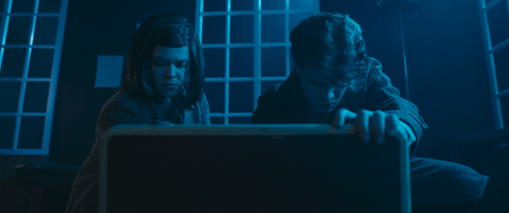 Two children, a girl and a boy, are leaning over and looking into a large, closed case or container, in a dimly lit room with blue lighting and shelves in the background.