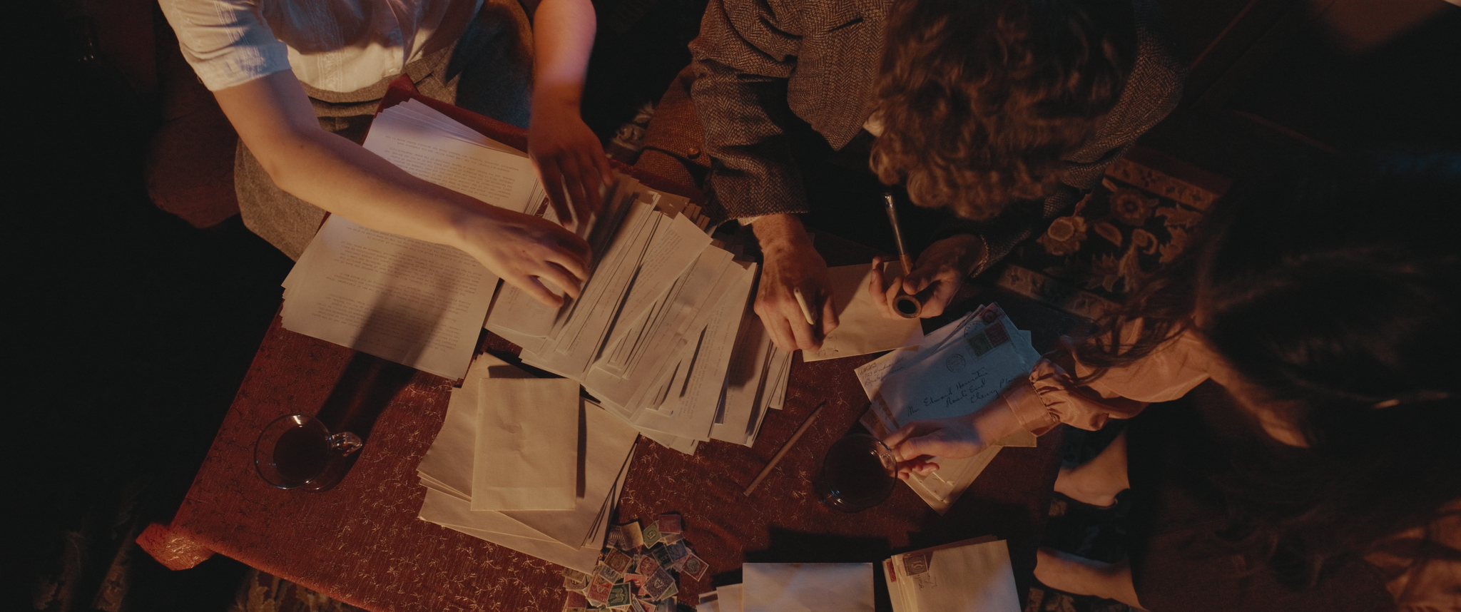Three women sitting around a table filled with papers, envelopes, and stamps, engaged in writing and sorting mail.