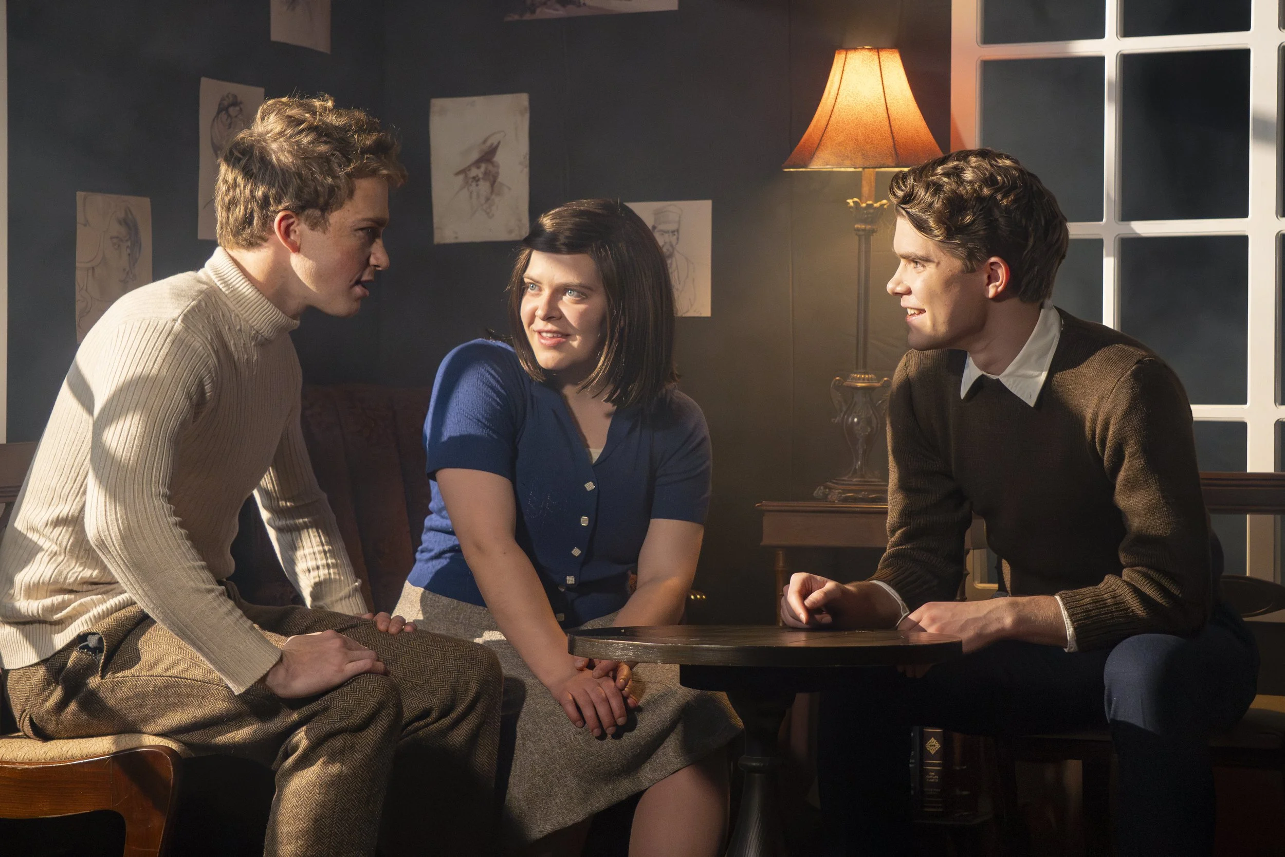 Three young adults sitting and engaging in conversation in a dimly lit room, with artwork on the wall and a table lamp providing warm lighting.