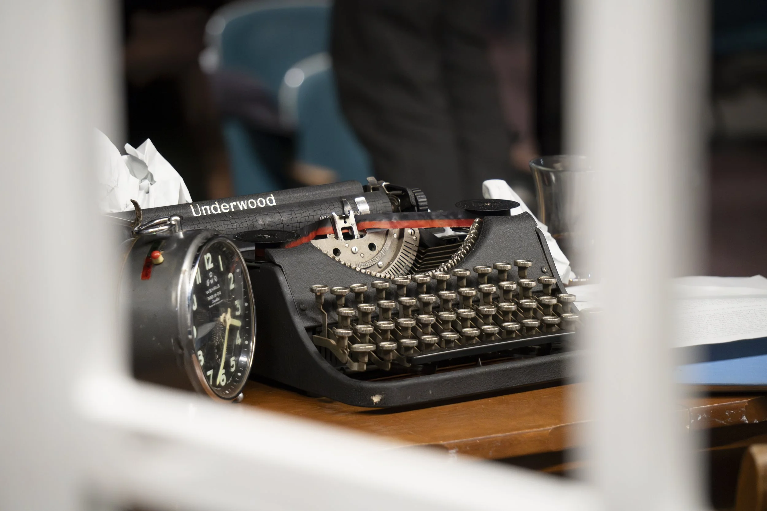 A vintage black typewriter with round keys on a wooden desk, an analog clock, a crumpled paper, a glass of water, and an open book, with a person blurred in the background.