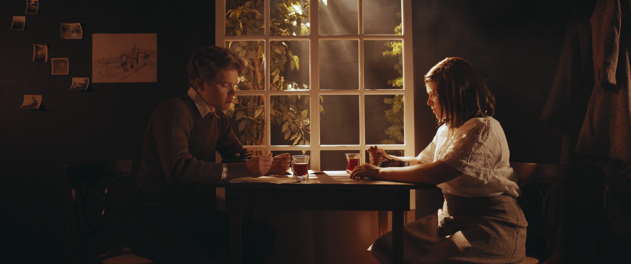 A young man and woman sitting at a table in a dimly lit room, having a meal with glasses of tea, with sunlight streaming through a large window behind them.