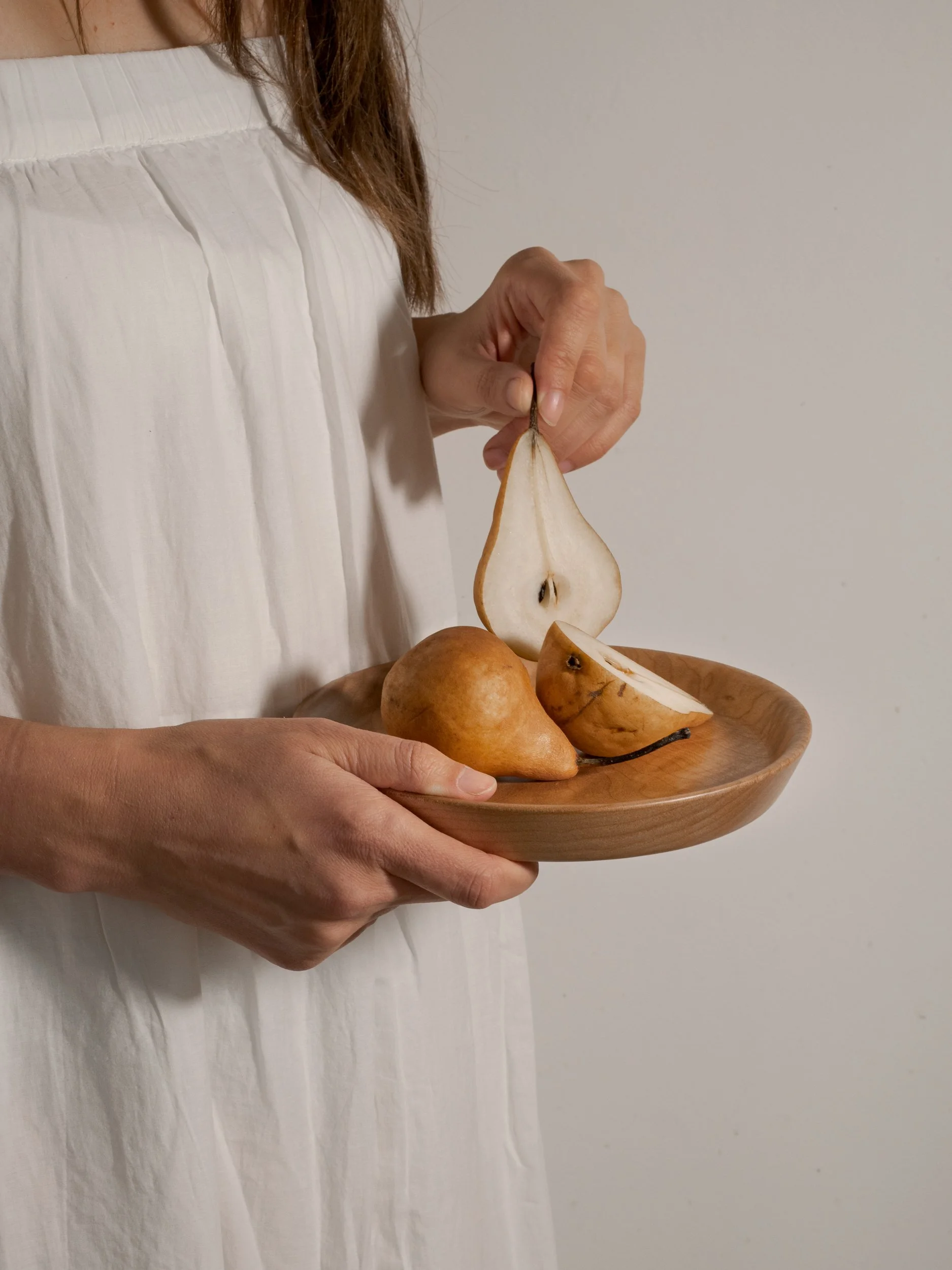 Person holding a wooden plate with sliced pears, including a pear slice hanging from their fingers. Product photography for wood worker and potter in Cowichan Valley.
