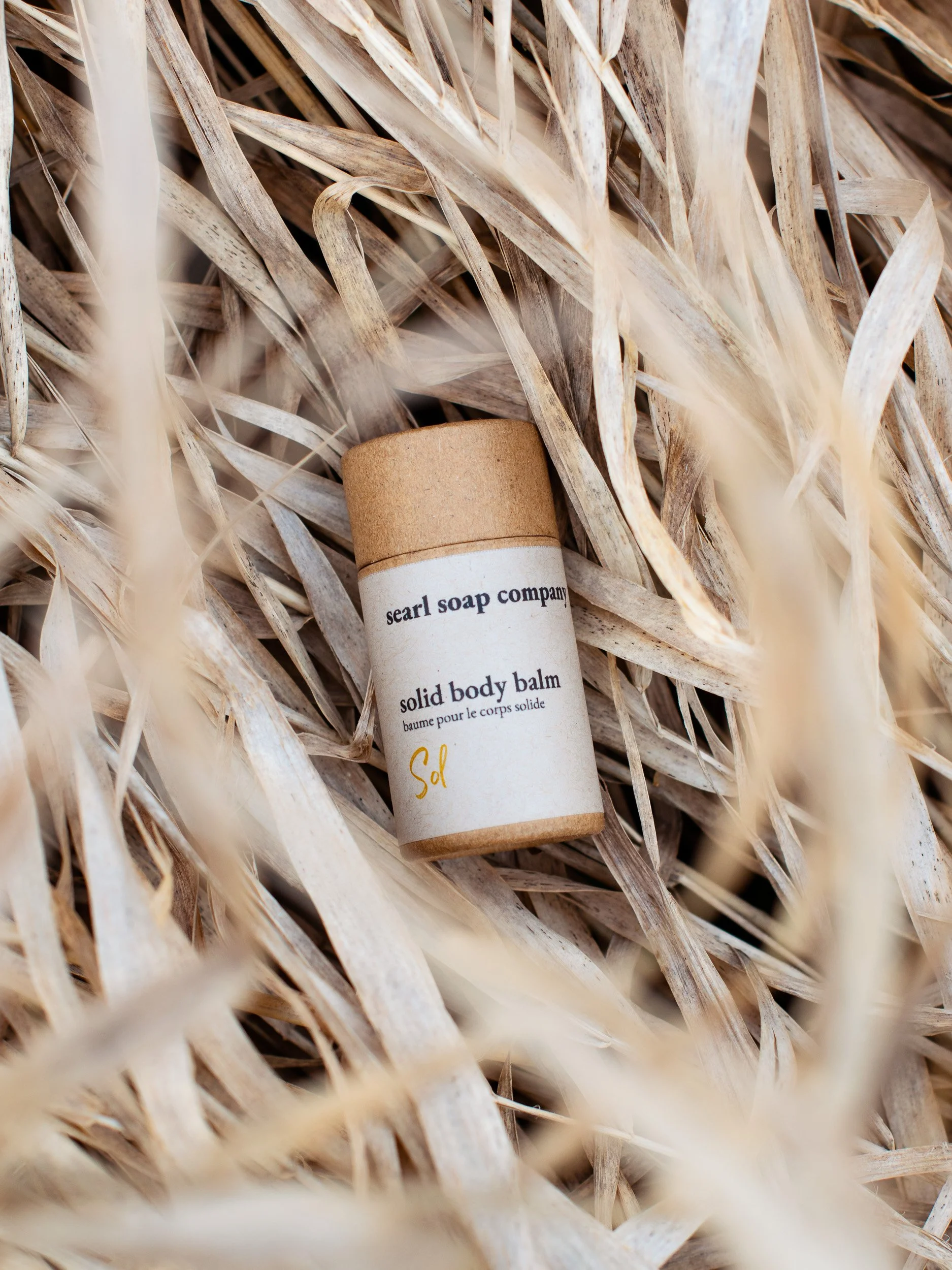 A small container of solid body balm from Searl Soap Company resting on dry, beige grass.