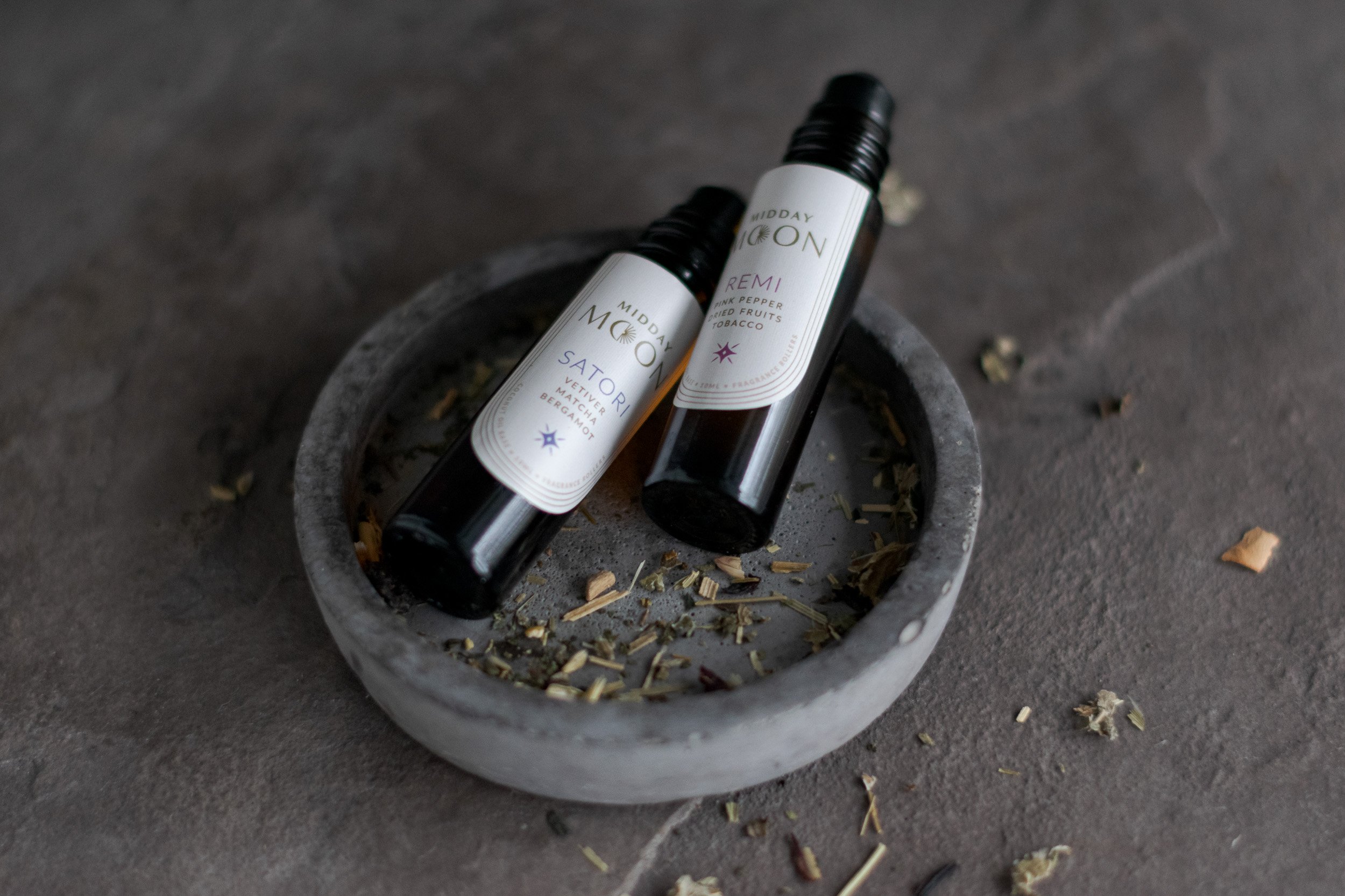 Two black bottles of scented oil resting in a round, gray stone bowl with dried herbs inside, placed on a gray surface.