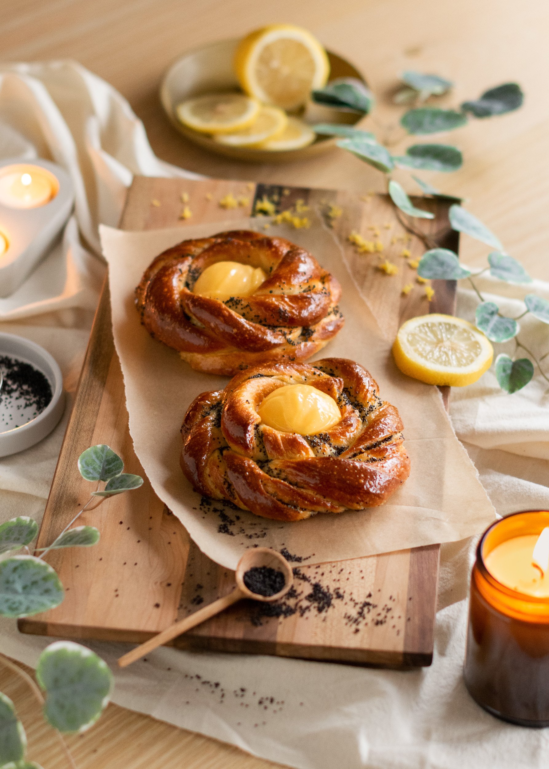 Two rose-shaped pastries with a yellow egg yolk in the center placed on parchment paper on a wooden board, surrounded by lemon slices, eucalyptus leaves, black poppy seeds, and tea lights.