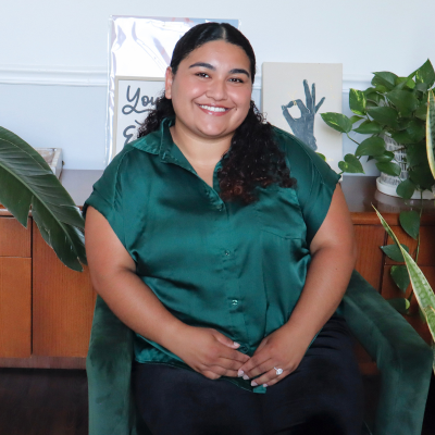 A smiling woman with dark hair, wearing a green satin blouse, sitting in a room with potted plants and decorative signs in the background.