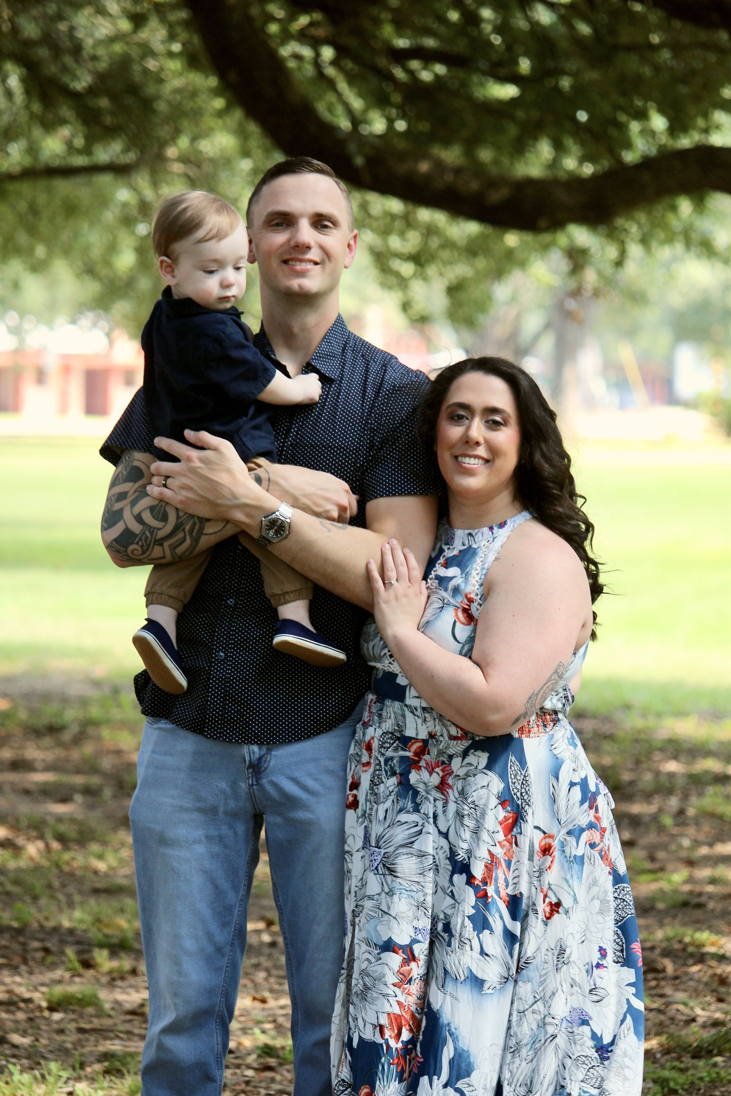 A family of three standing outdoors under a large tree, smiling at the camera. The man is holding a young child, and the woman is standing beside them.