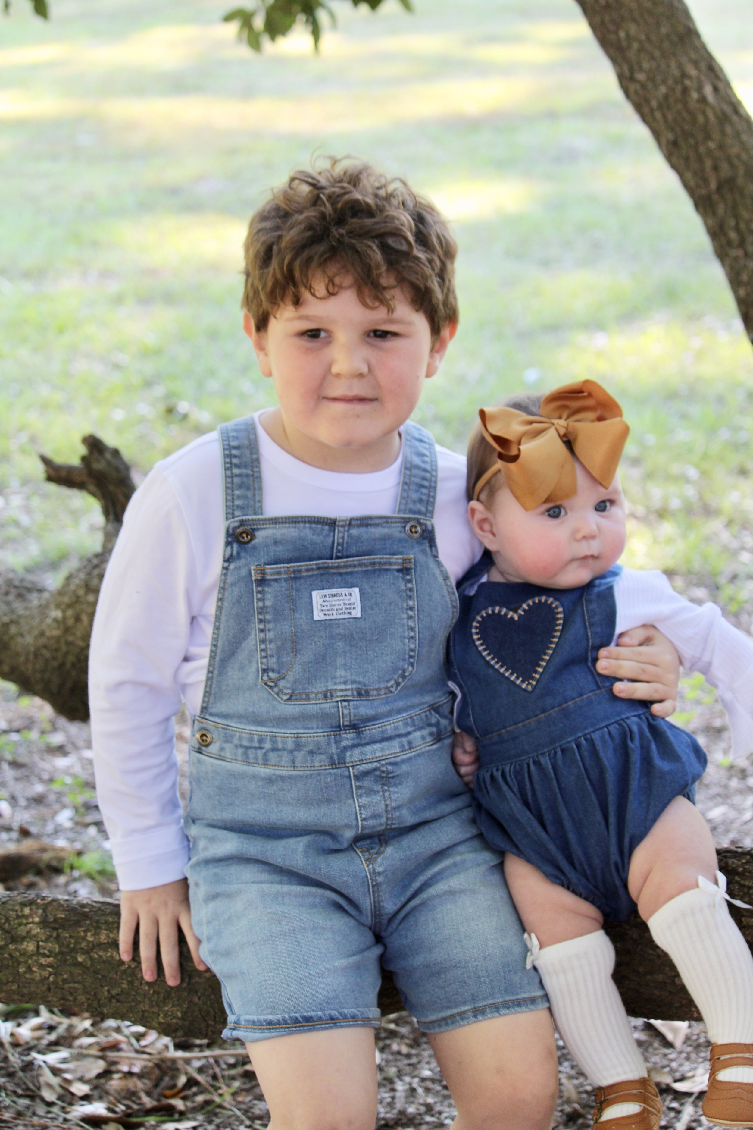 A young boy with curly brown hair and a white shirt in denim overalls sitting on a fallen tree trunk outdoors with a younger girl in a blue dress with a large bow on her head and white knee-high socks, under a shaded tree.
