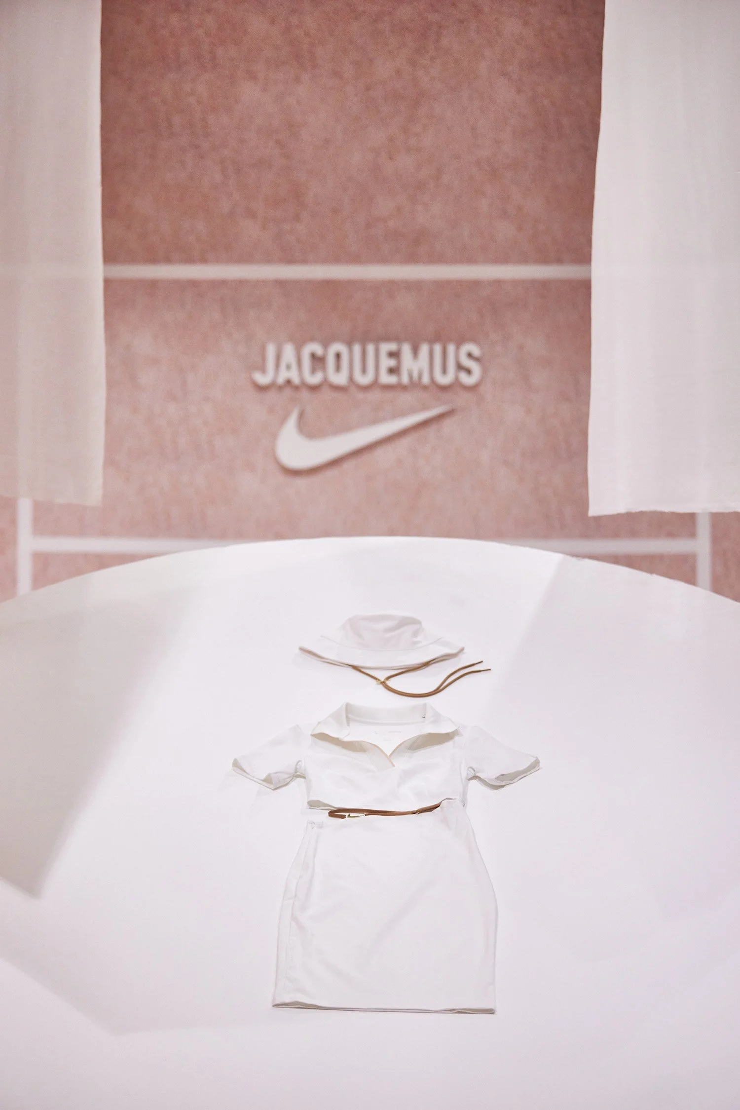 A white dress, hat with brown string, and belt arranged on a white table, with a pink tiled wall and "JACQUEMUS" and Nike logo in the background.