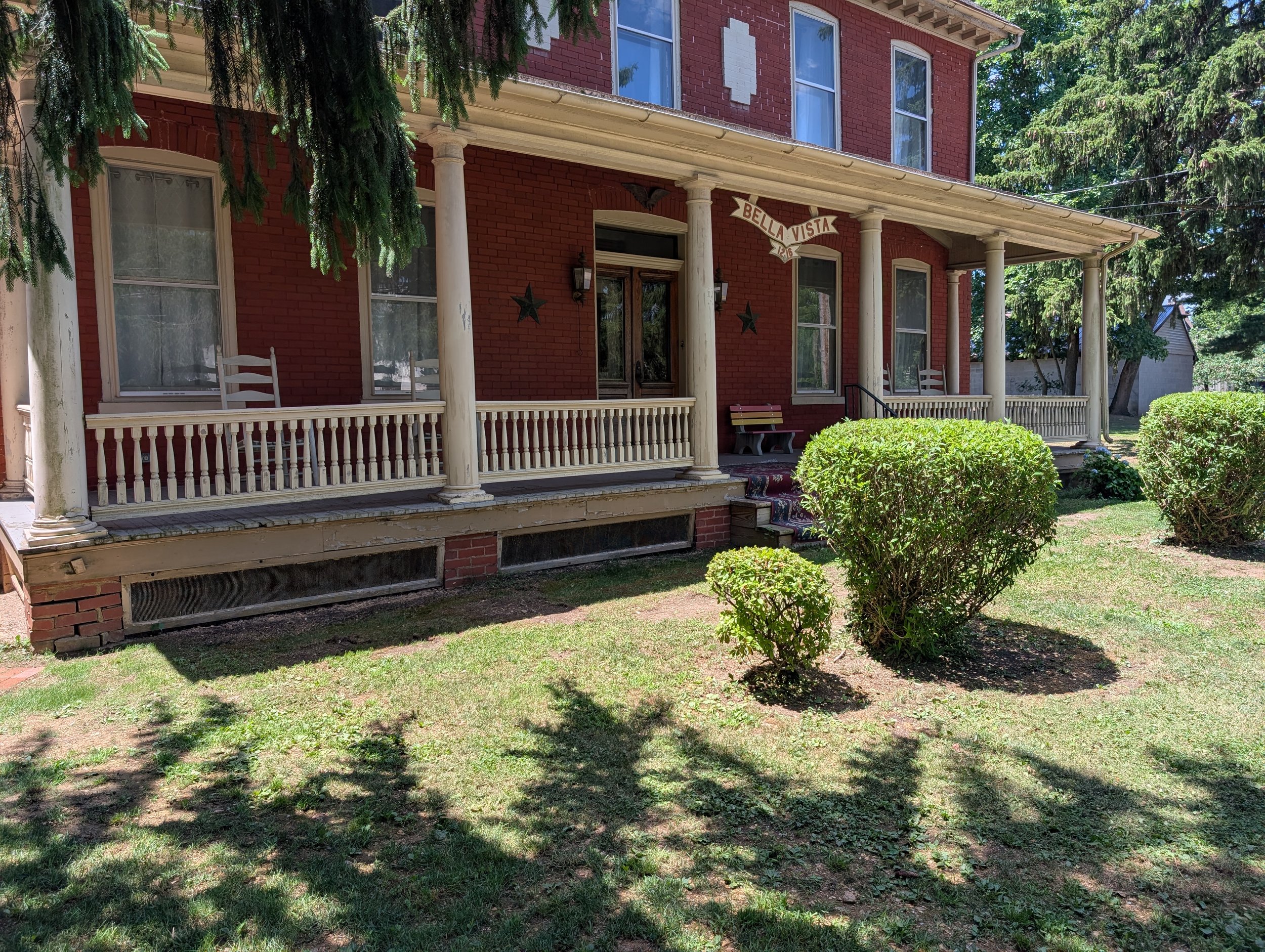 Welcoming front porch at Bella Vista Bed and Breakfast, surrounded by landscaped gardens and shaded by mature trees in historic Akron, Pennsylvania.