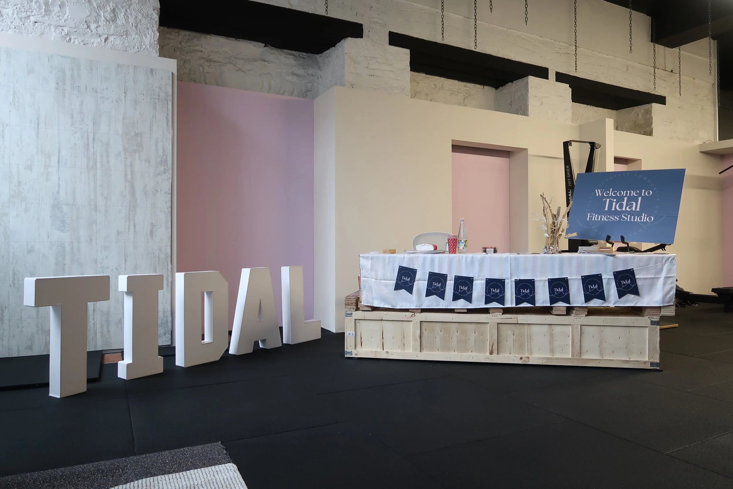 Reception desk with a sign that reads 'Welcome to Tidal Fitness Studio,' pink and white walls, large white letters spelling 'TIDAL,' and fitness/gym equipment in the background.