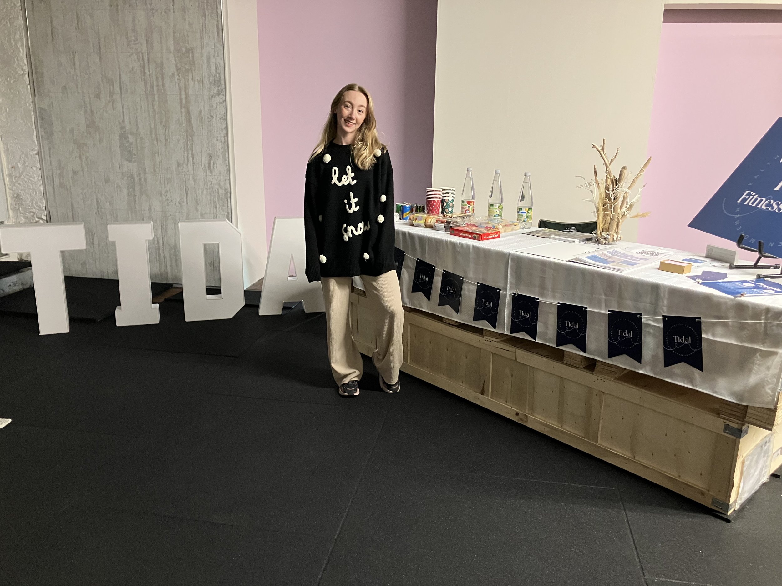 The friendly and approachable owner of Tidal Fitness standing next to a table decorated for an event, with large white letters spelling 'TIDAL' behind her and event materials on the table.