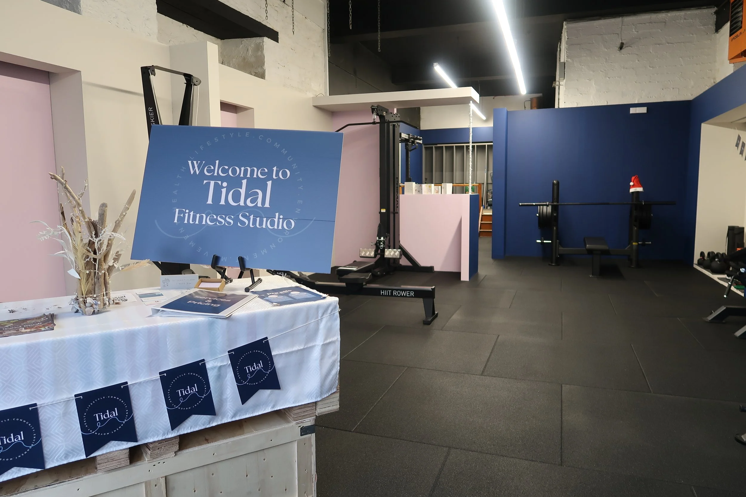 Interior of a fitness studio/gym with a sign reading 'Welcome to Tidal Fitness Studio'. There is a table with information brochures and decorations, a lat pull-down machine, a weight bench, and a wall with different workout equipment.