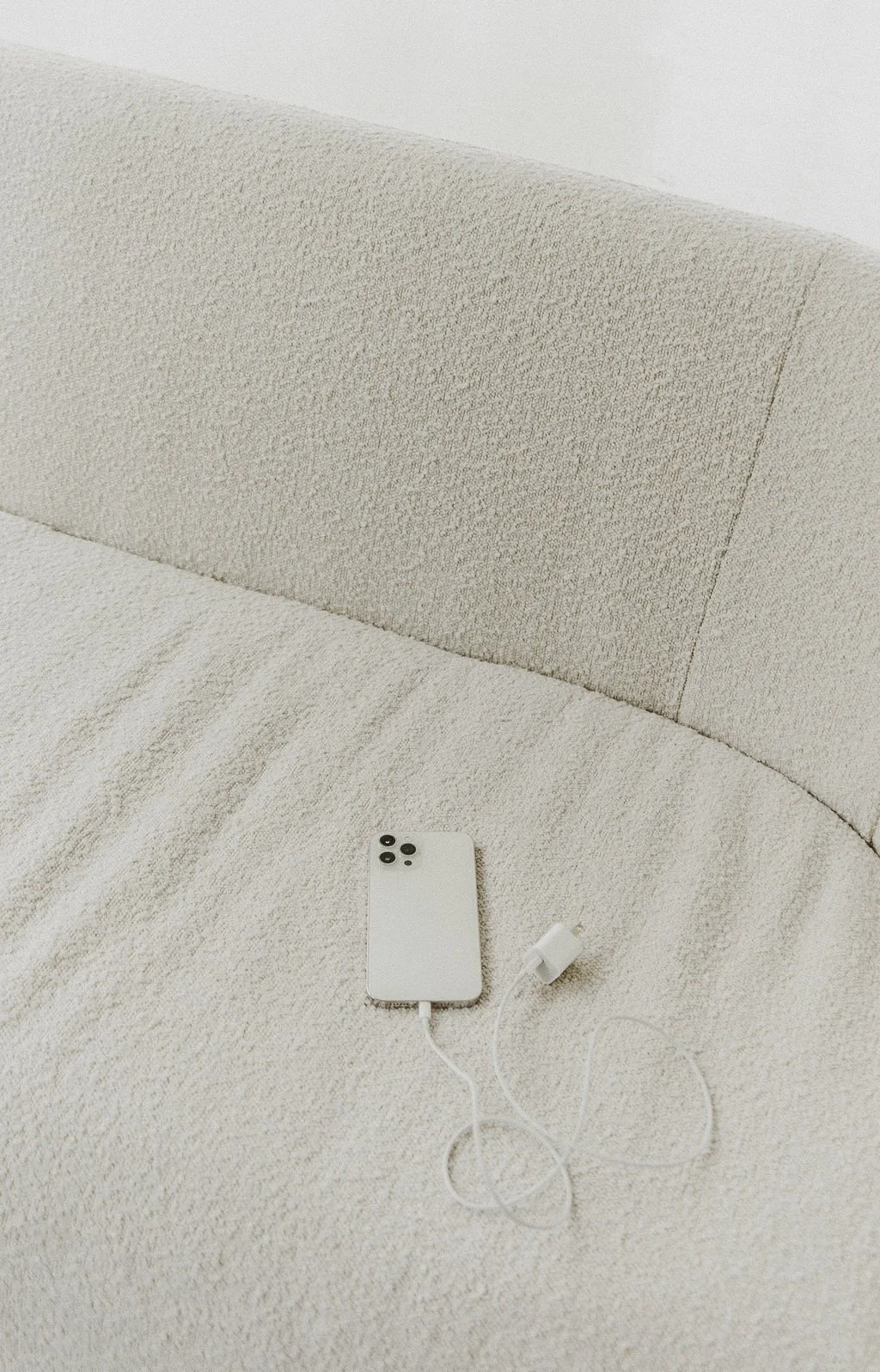 Smartphone with a white case and charging cable lying on a cream-colored textured fabric surface, possibly a sofa or bed.