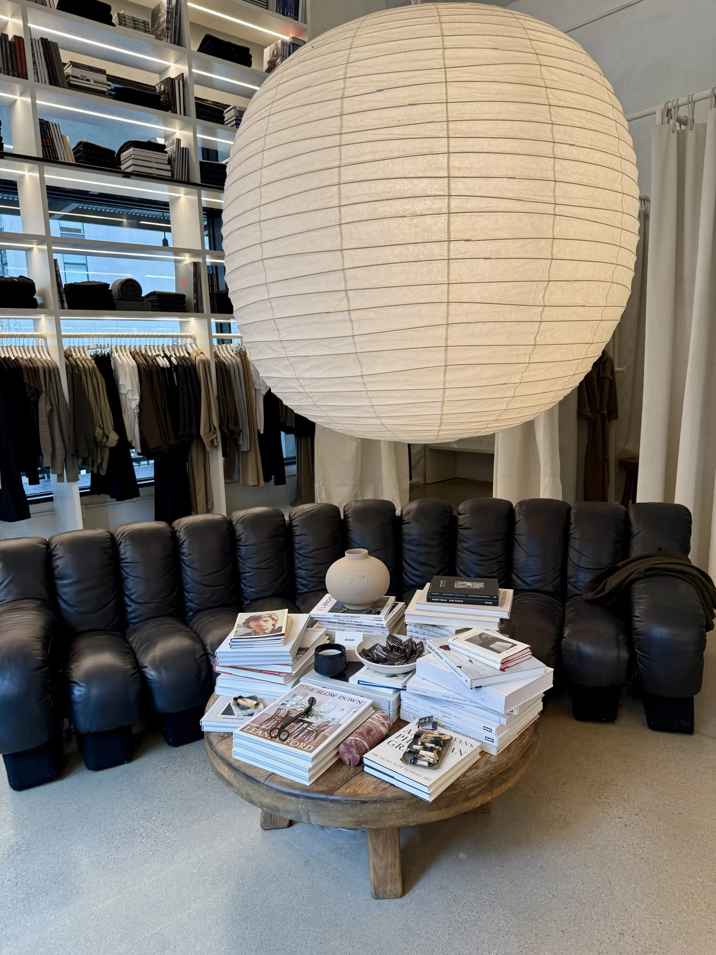 A round wooden coffee table covered with stacks of books and magazines, with a small white vase and some decorative items on top, in a living room with a black leather sofa, a large paper lantern, and a bookshelf with clothing and books in the background.