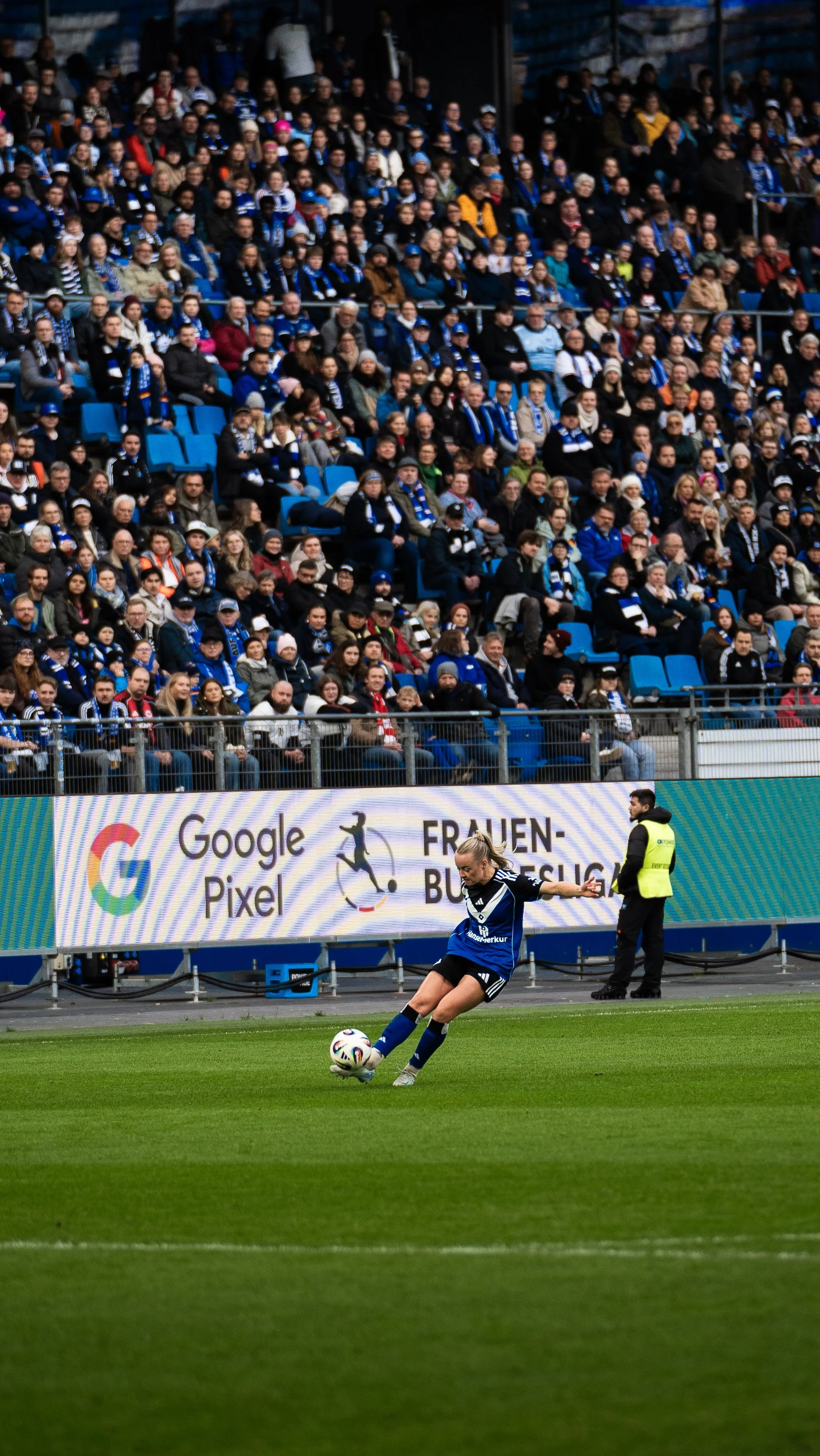 Fußballspiel, weibliche Spielerin bei einem Freistoß auf einem grünen Rasen, viele Zuschauer im Hintergrund, IT-Audience trägt blau-weißes Halstuch, digitaler Bildschirm im Hintergrund mit Google Pixel und Frauen-Büros-Liga