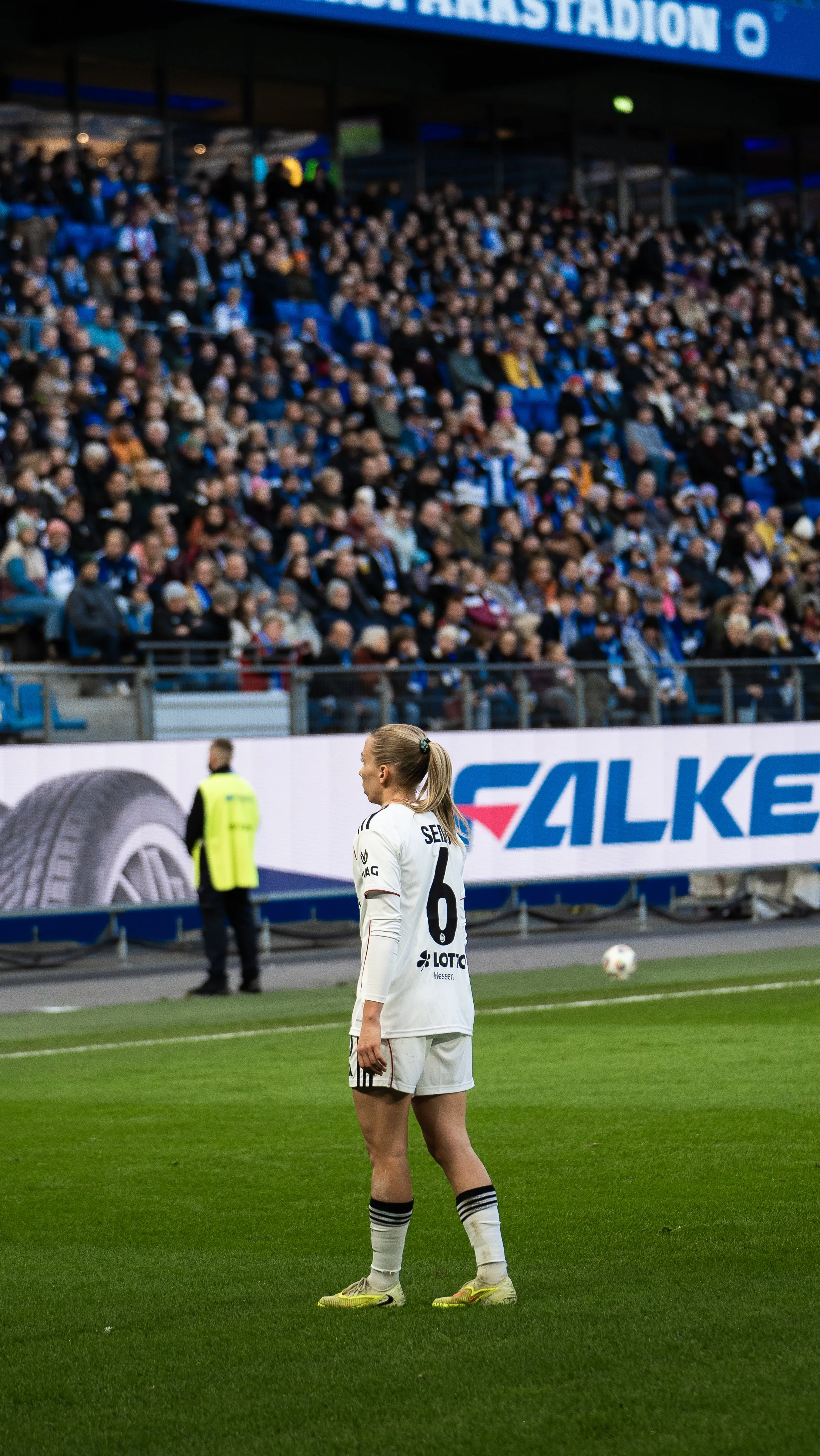 Fußballspielerin in weißem Trikot und kurzen Shorts auf dem Spielfeld mit einer vollen Stadionkulisse im Hintergrund.