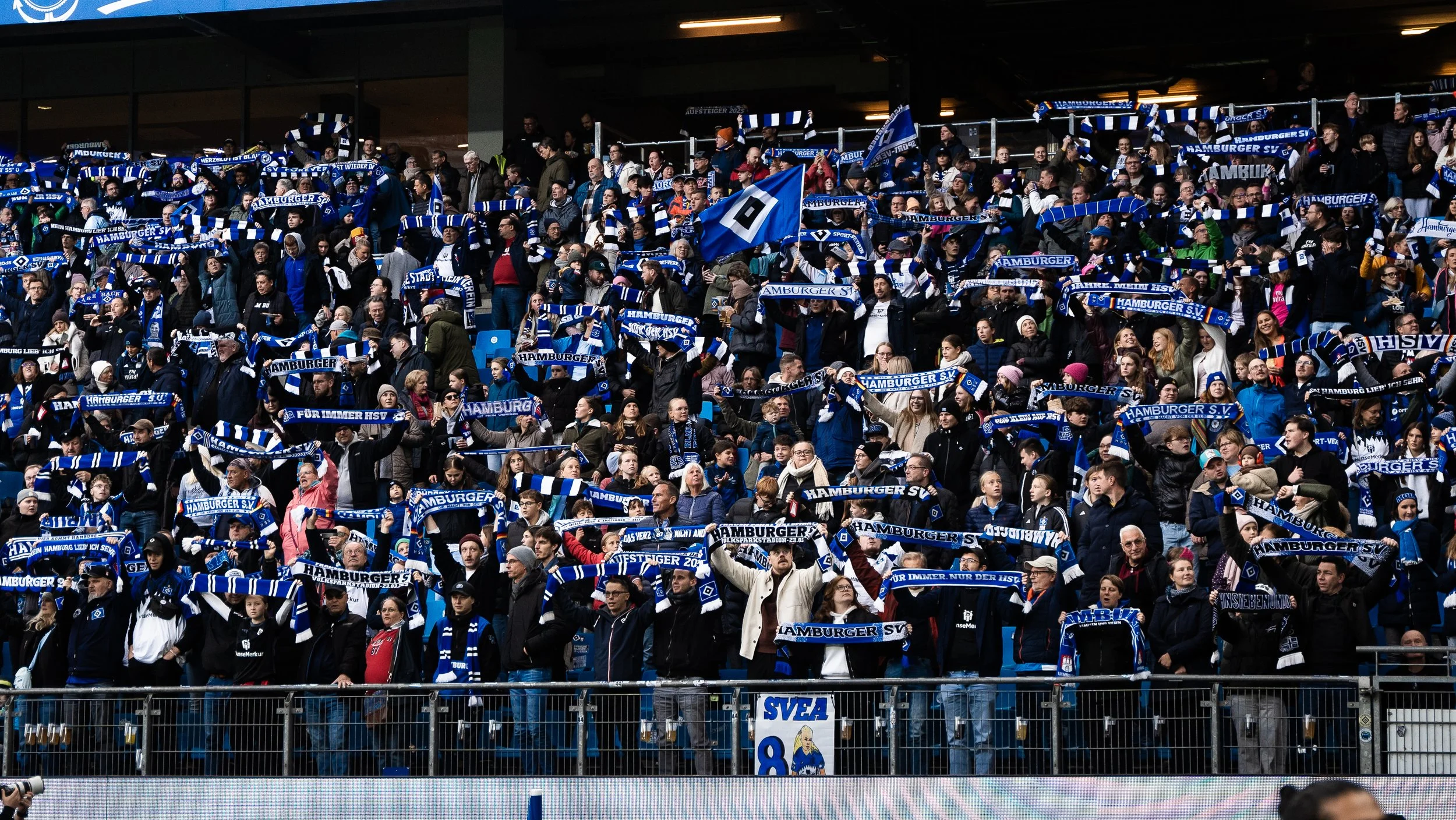 Menschenmenge auf einem Stadiontribüne, die den HSV-Fanclub unterstützt, mit blauen und weißen Schals und Fahnen, die HSV-logos zeigen.