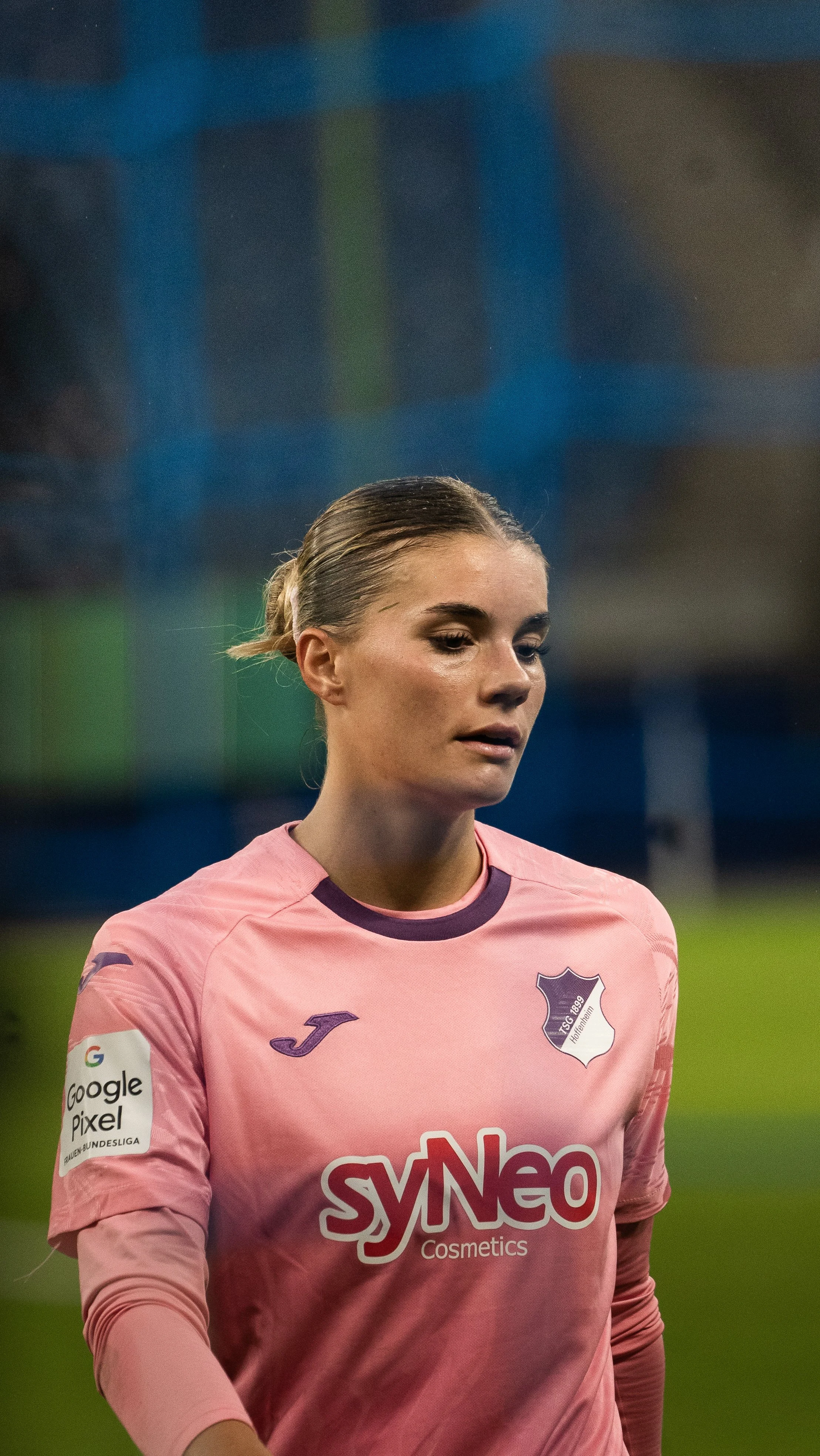 Eine blonde Fussballspielerin trägt ein pinkfarbenes Trikot mit dem Logo der TSG Hoffenheim und schaut nach unten auf einem Fußballfeld.