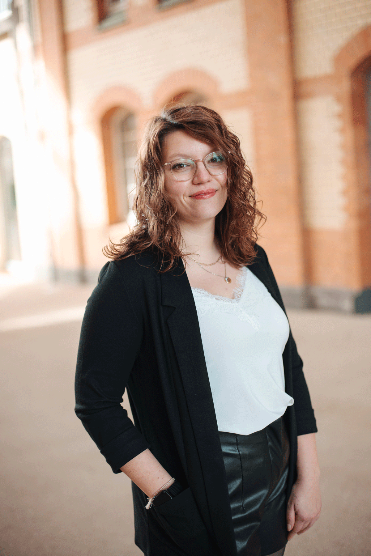 Junge Frau mit lockigem, braunem Haar, trägt Brille, dunkle Jacke, weiße Bluse und Lederhosen, steht vor einer Backsteinwand im Freien.