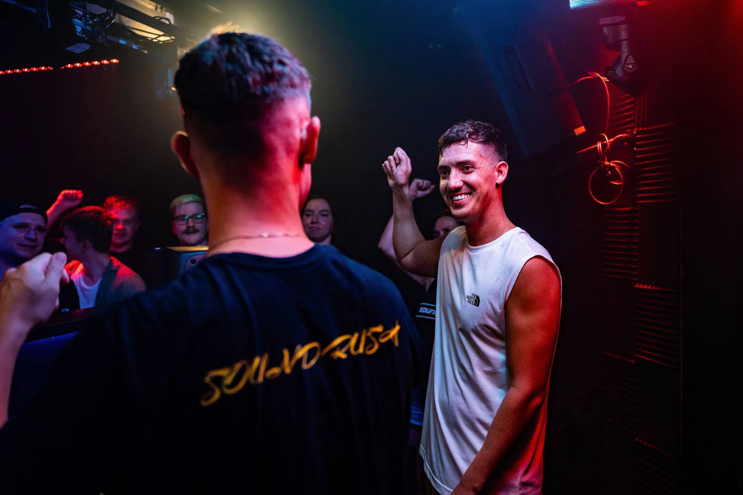Zwei Männer in einem Club, einer mit einem weißen Tanktop und der andere mit einem schwarzen T-Shirt, lachen und machen eine Geste der Stärke, während sie von anderen Menschen im Hintergrund beobachtet werden.