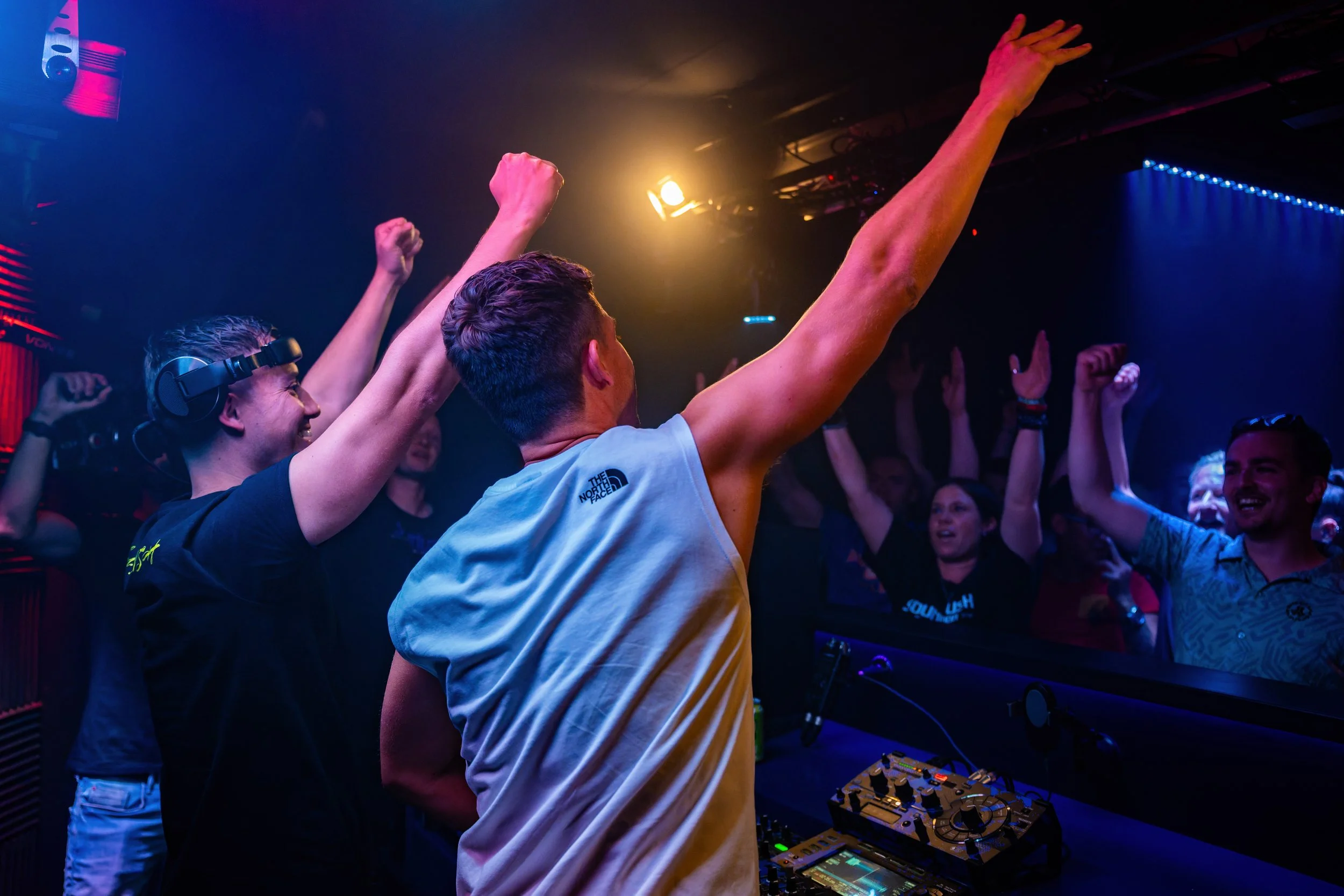 Menschen feiern in einem Club mit DJ, tanzen und heben die Arme in die Luft.