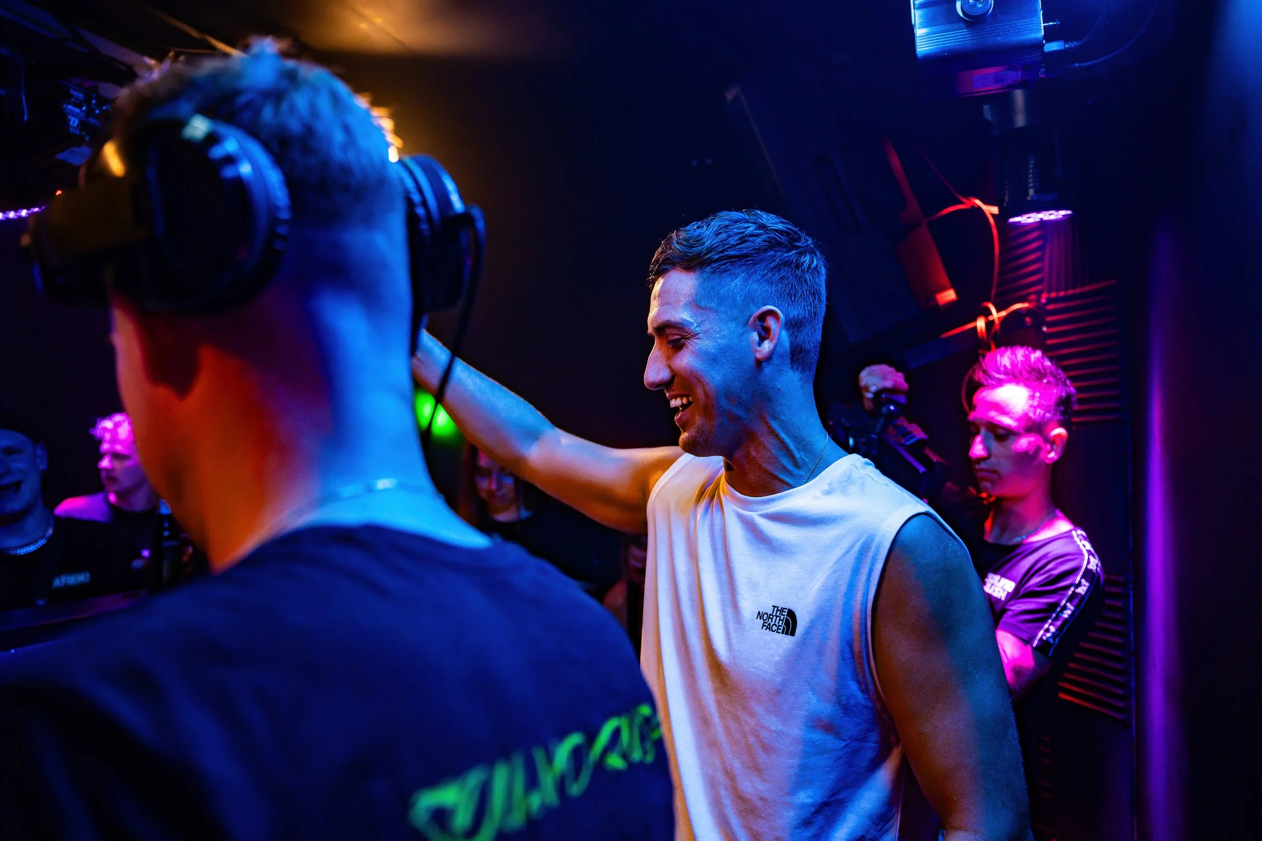 Menschen beim Feiern in einem dunklen Club, bunte Neonlichter, einer trägt ein weißes T-Shirt von The North Face, lächelt, andere tragen Kopfhörer.