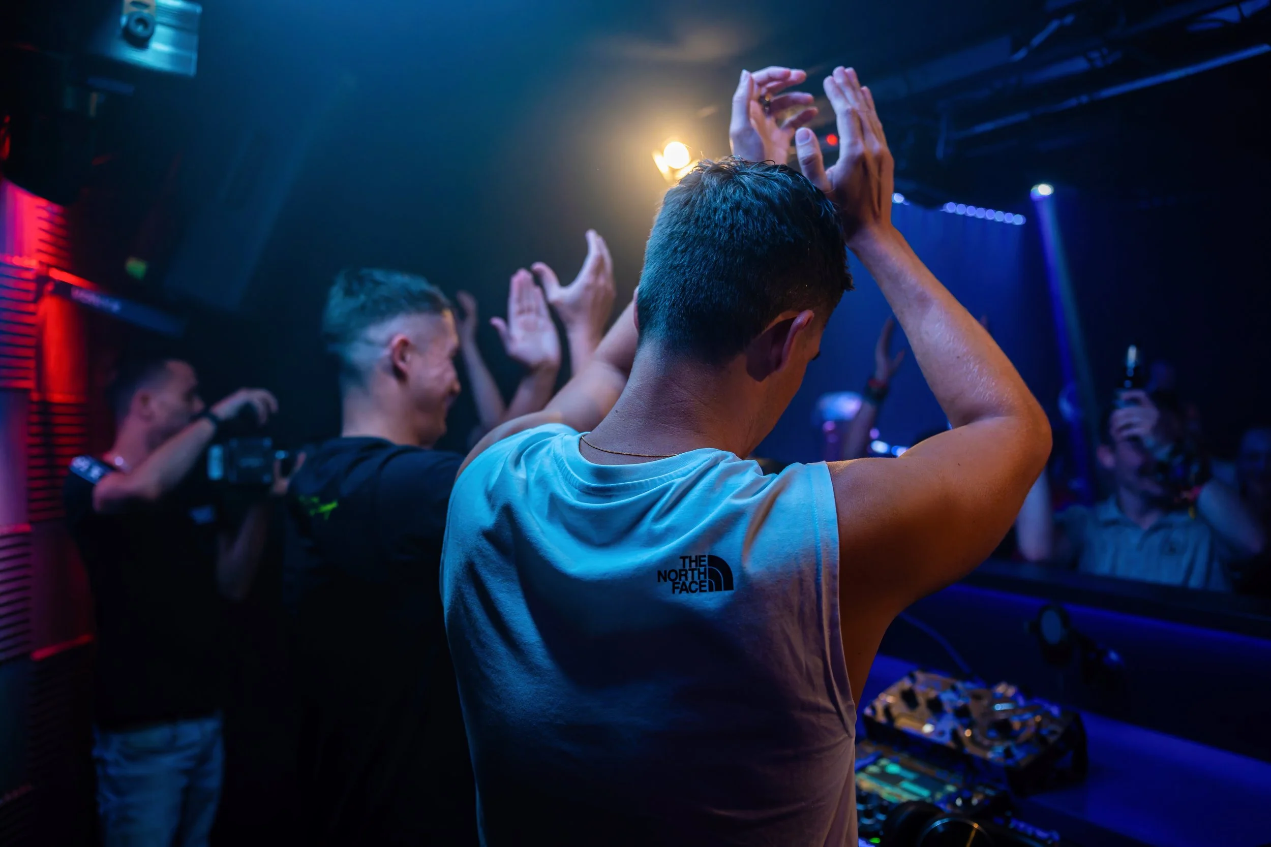 Menschen feiern in einem Club, tanzen zur Musik, einige haben Getränke in der Hand, im Vordergrund ein junger Mann mit Kurzhaarschnitt und Tanktop von The North Face, im Hintergrund bunte Lichter und DJ-Equipment.