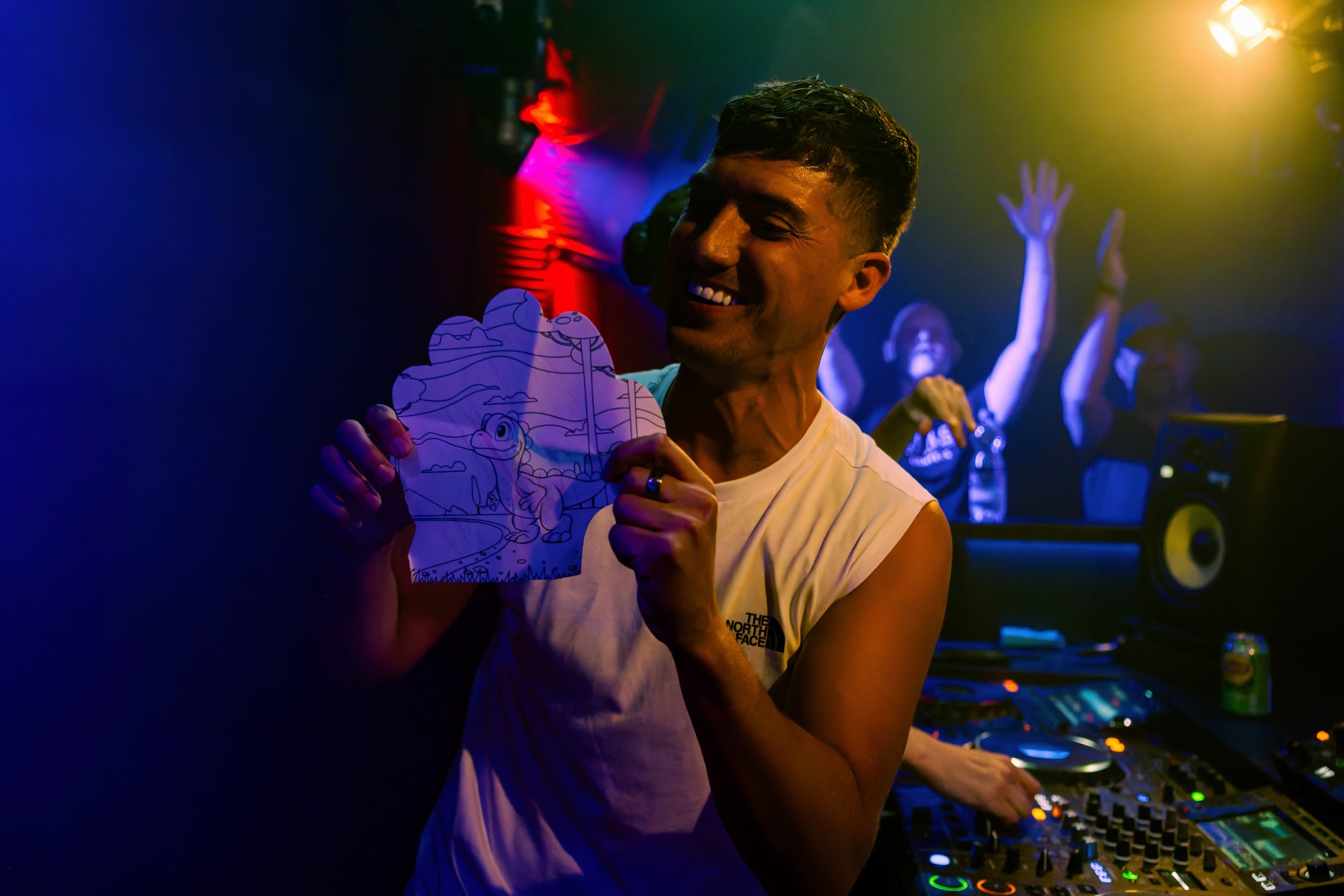 Ein lachender Mann in einem Fitness-Shirt hält ein handgemaltes Papierbild mit Drachenszenen in einer Party- oder Club-Umgebung, im Hintergrund tanzen Menschen unter bunten Lichteffekten.