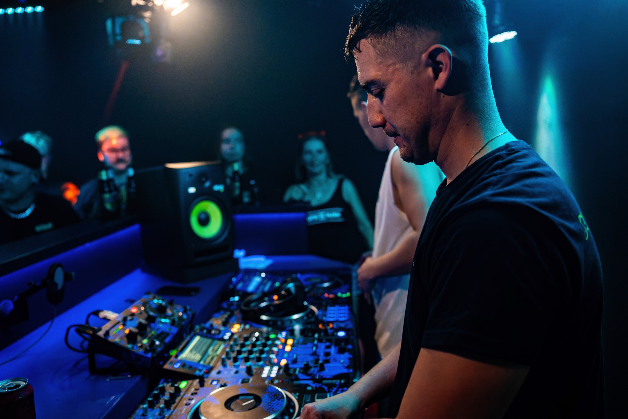 DJ spielt auf einer Party mit Musik equipment, umgeben von feiernden Gästen im dunklen Club, mit blauen und grünen Lichteffekten.